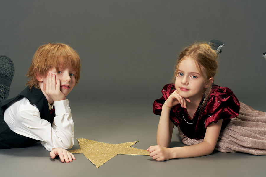 A boy and a girl lie on the floor with a large gold star between them; the girl wears the KIKI and JOJO Girls Velvet Puff Sleeve Dress with Rhinestone Trim in Burgundy Red, both looking at the camera against a plain gray background.