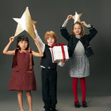 Three children pose against a gray backdrop; one wears the KIKI and JOJO Girls Handmade Tweed Dress with a Burgundy Velvet Bow, while two hold gold star props. The center child in this dress holds a white gift box with a red ribbon.