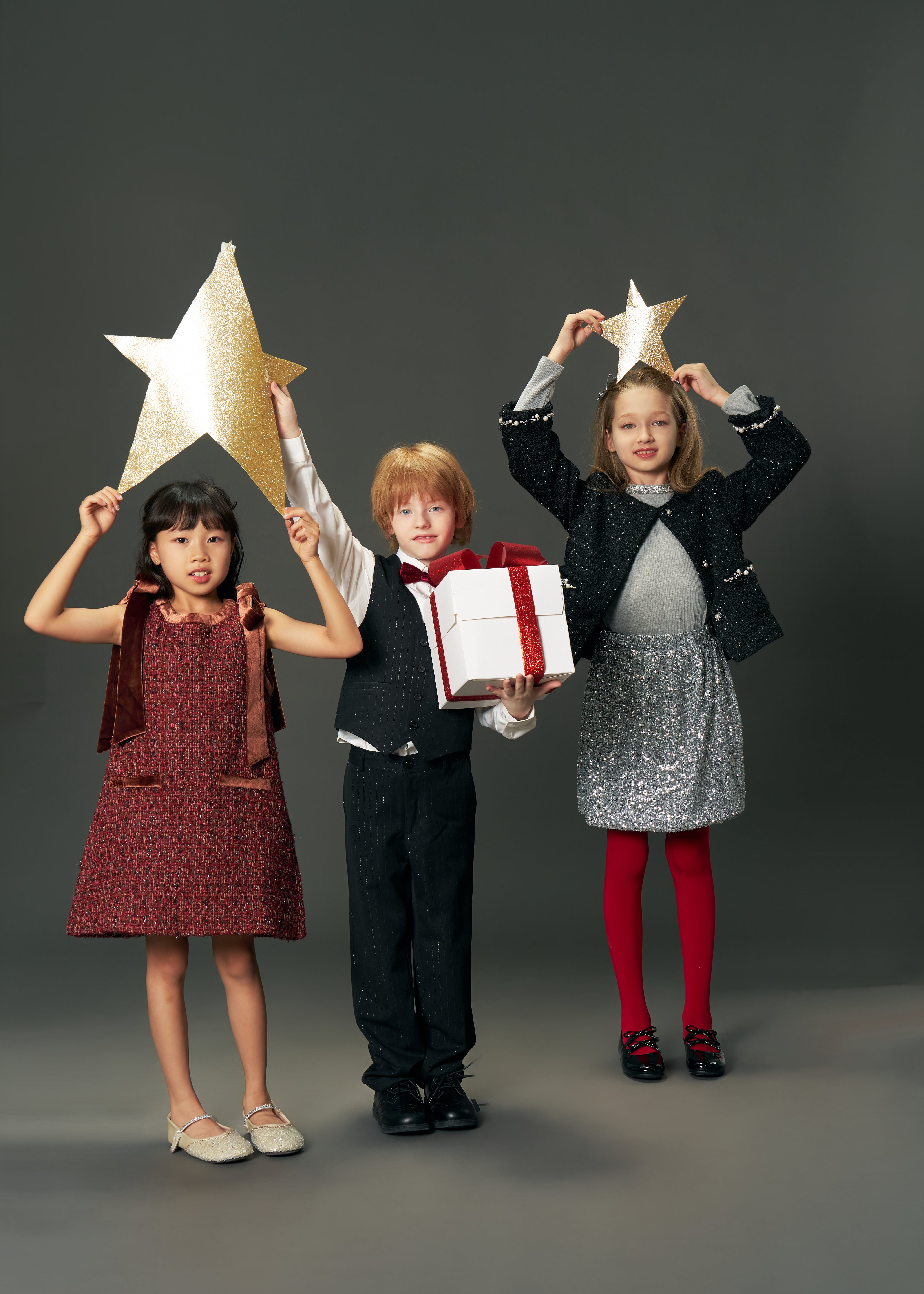 Three children pose against a gray backdrop; one wears the KIKI and JOJO Girls Handmade Tweed Dress with a Burgundy Velvet Bow, while two hold gold star props. The center child in this dress holds a white gift box with a red ribbon.