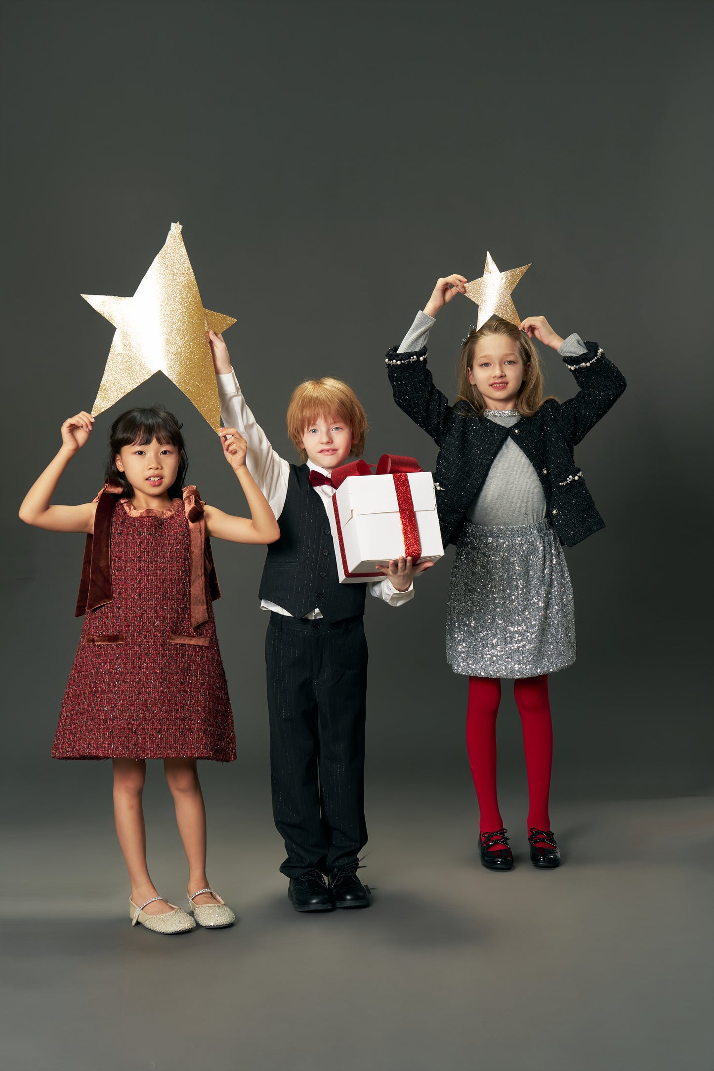 Three children pose against a gray backdrop; one wears the KIKI and JOJO Girls Handmade Tweed Dress with a Burgundy Velvet Bow, while two hold gold star props. The center child in this dress holds a white gift box with a red ribbon.