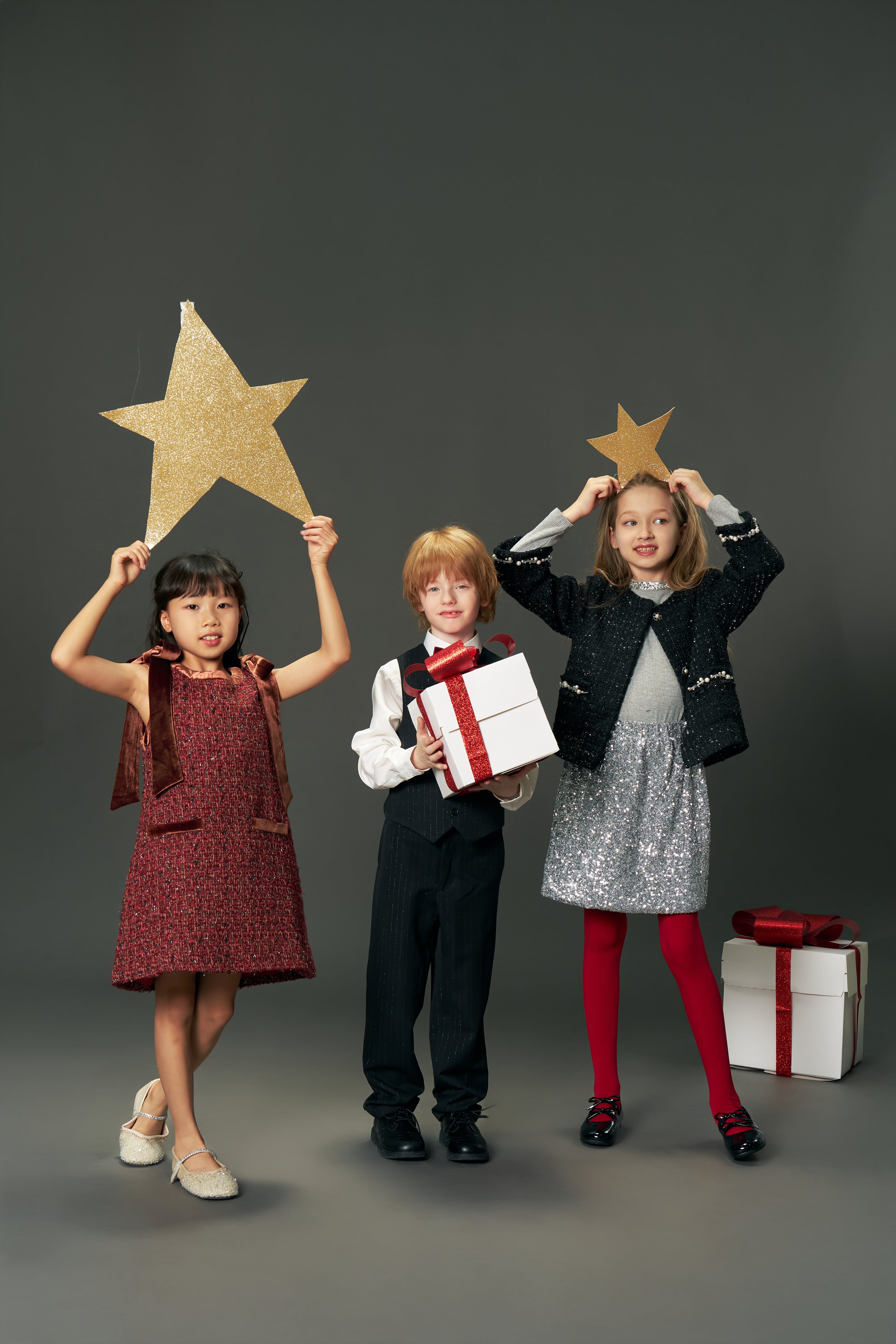 Three children in festive outfits pose in a studio. One wears the KIKI and JOJO Girls Handmade Tweed Dress – Burgundy Velvet Bow. Two hold gold star decorations, one holds a wrapped gift, and another gift box rests on the floor beside them.
