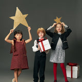 Three children in festive attire pose together; one wears the KIKI and JOJO Girls Tweed Jacket – Classic Black Pearl, while two hold gold stars overhead and another holds a white gift box with a red ribbon. Another gift box sits on the floor nearby.