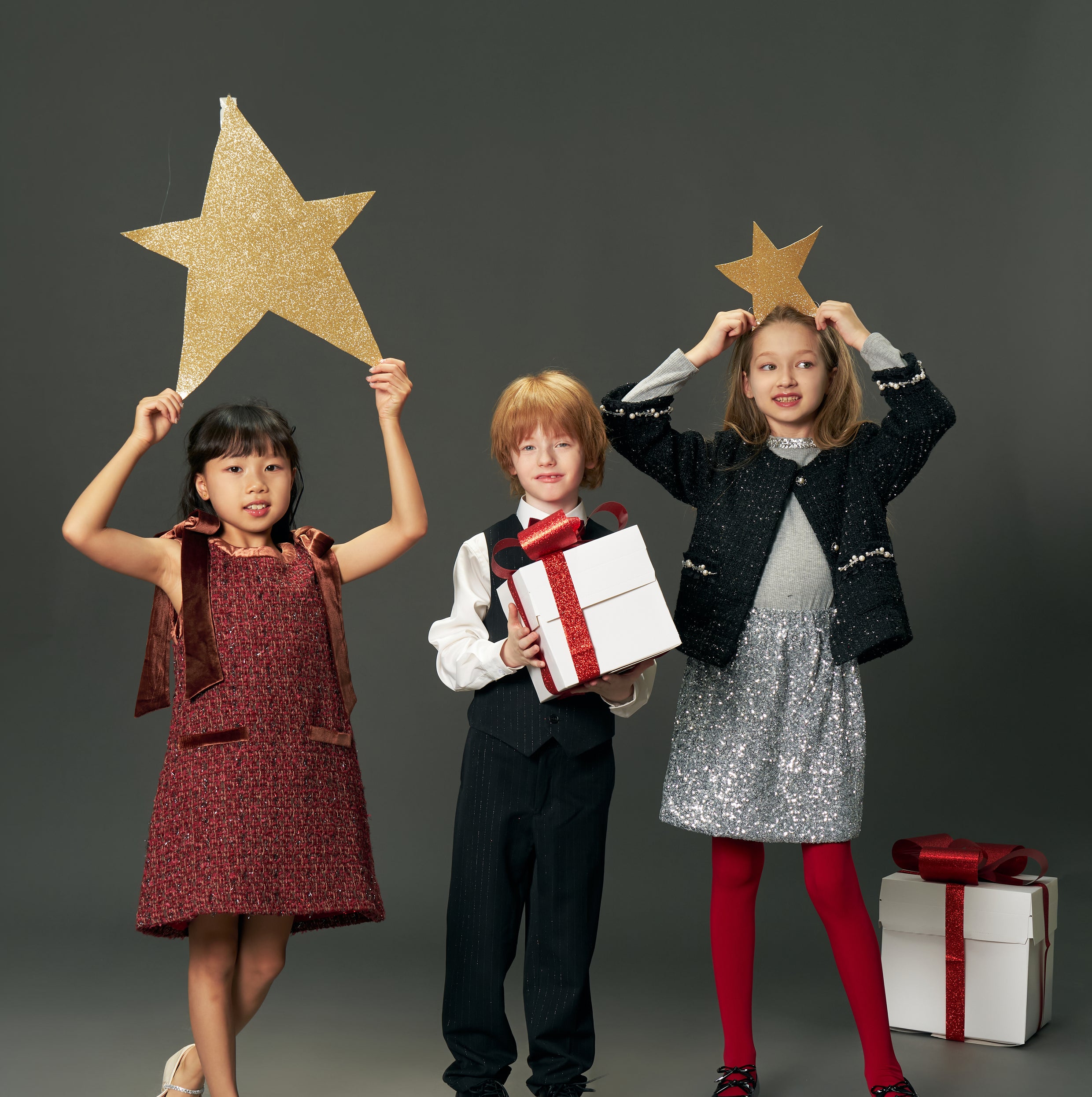 Three children in festive attire pose together; one wears the KIKI and JOJO Girls Tweed Jacket – Classic Black Pearl, while two hold gold stars overhead and another holds a white gift box with a red ribbon. Another gift box sits on the floor nearby.
