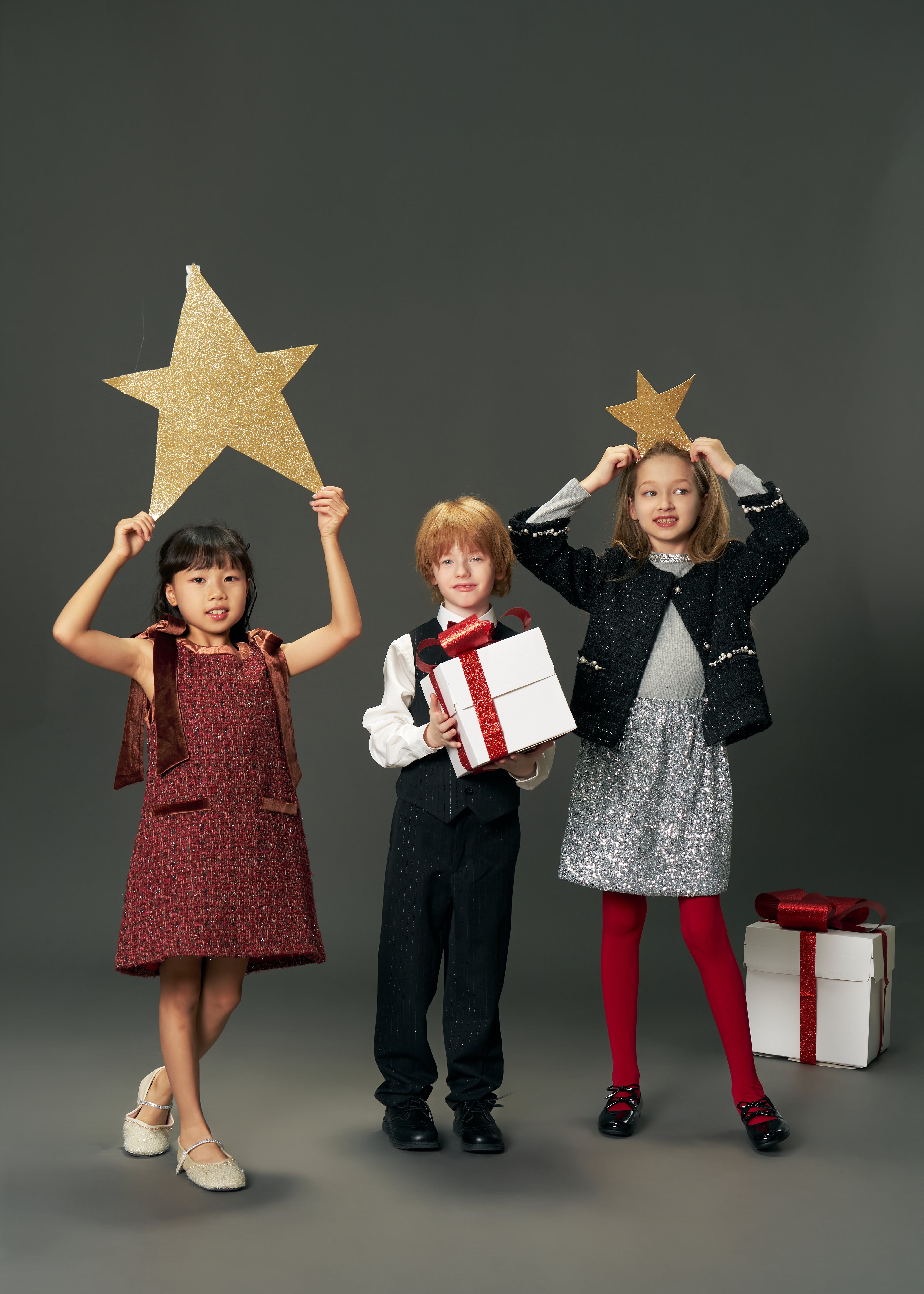 Three children in festive attire pose together; one wears the KIKI and JOJO Girls Tweed Jacket – Classic Black Pearl, while two hold gold stars overhead and another holds a white gift box with a red ribbon. Another gift box sits on the floor nearby.