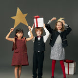 Three children in festive outfits stand side by side holding gold stars and gift boxes; one girl wears the KIKI and JOJO Girls Handmade Tweed Dress with a burgundy velvet bow, set against a plain gray background.