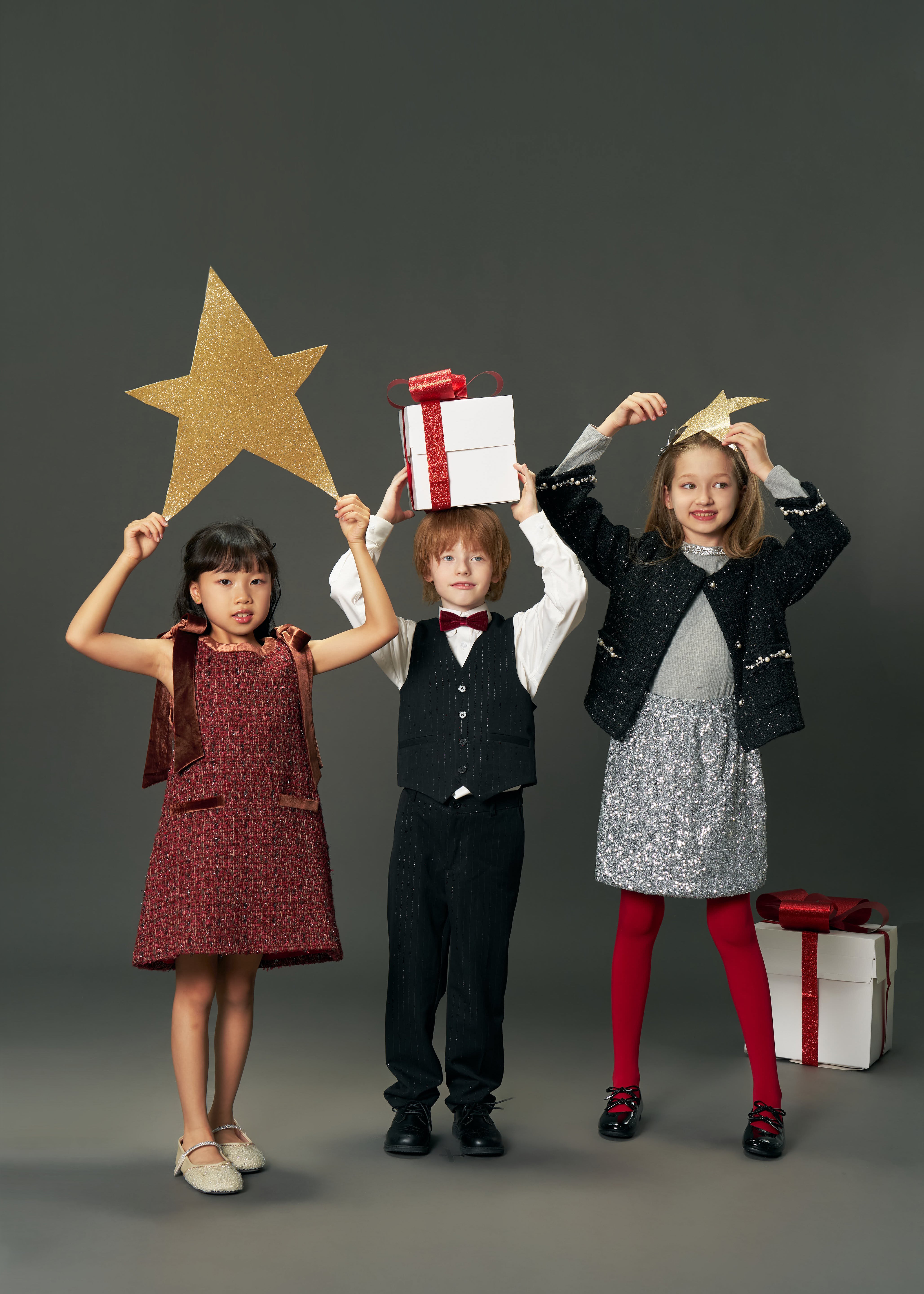 Three children in festive outfits stand side by side holding gold stars and gift boxes; one girl wears the KIKI and JOJO Girls Handmade Tweed Dress with a burgundy velvet bow, set against a plain gray background.
