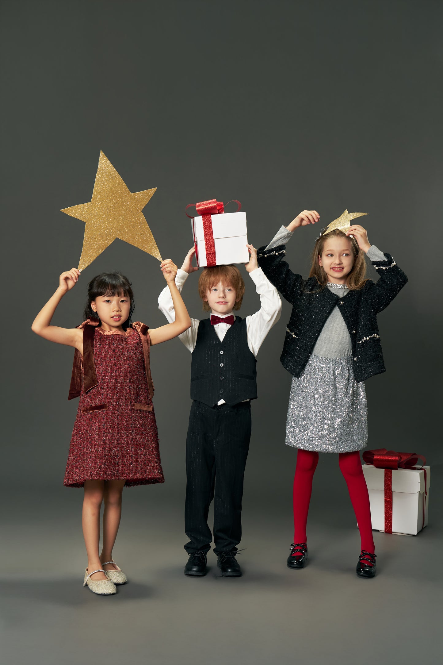 Three children in festive outfits stand side by side holding gold stars and gift boxes; one girl wears the KIKI and JOJO Girls Handmade Tweed Dress with a burgundy velvet bow, set against a plain gray background.
