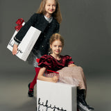 Two young girls pose festively; one holds a gift bag, while the other sits in a box labeled "kikiyaja," wearing the KIKI and JOJO Girls Tweed Jacket – Classic Black Pearl with pearl details, set against a plain gray background.
