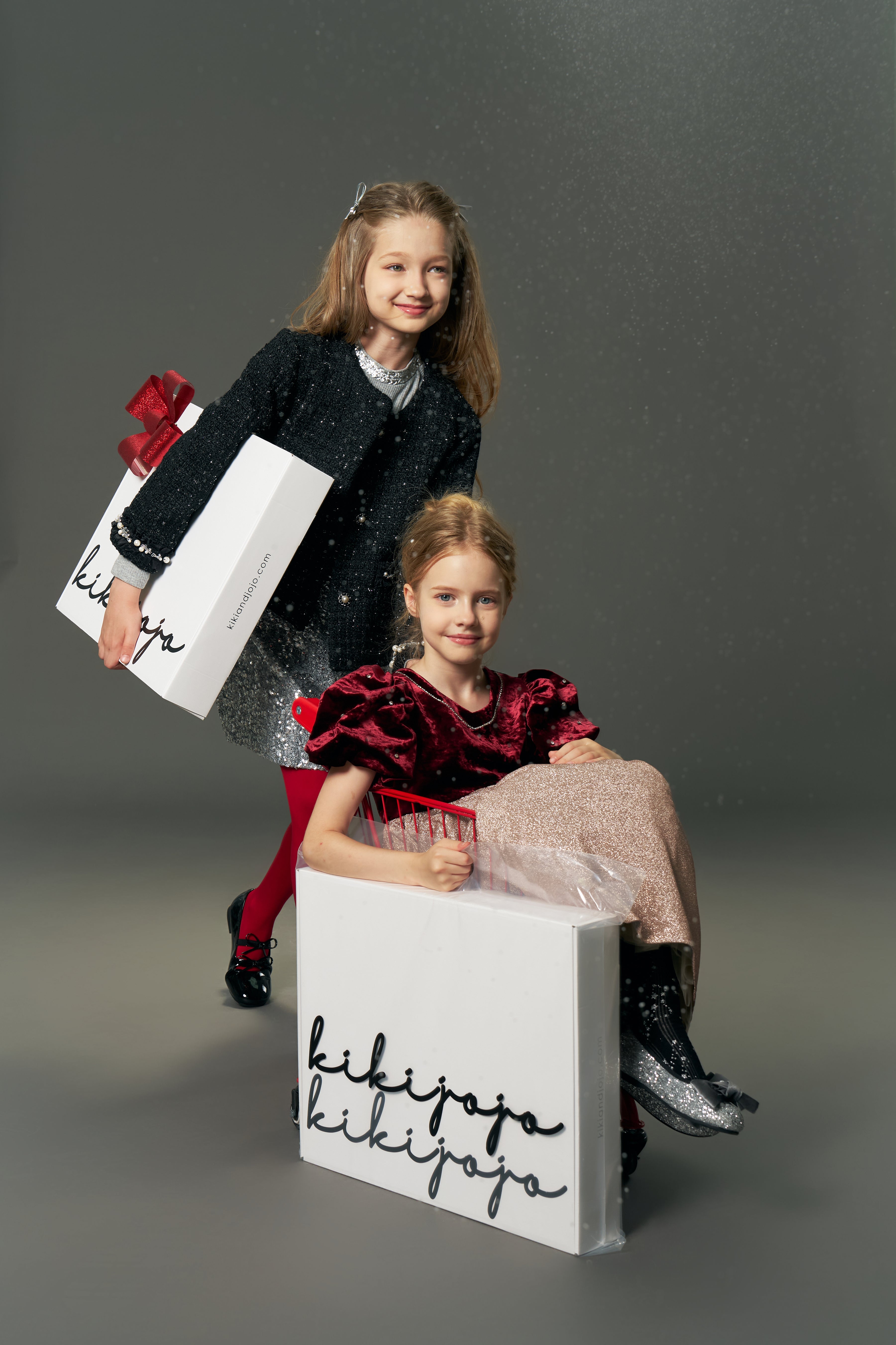 Two young girls pose festively; one holds a gift bag, while the other sits in a box labeled "kikiyaja," wearing the KIKI and JOJO Girls Tweed Jacket – Classic Black Pearl with pearl details, set against a plain gray background.