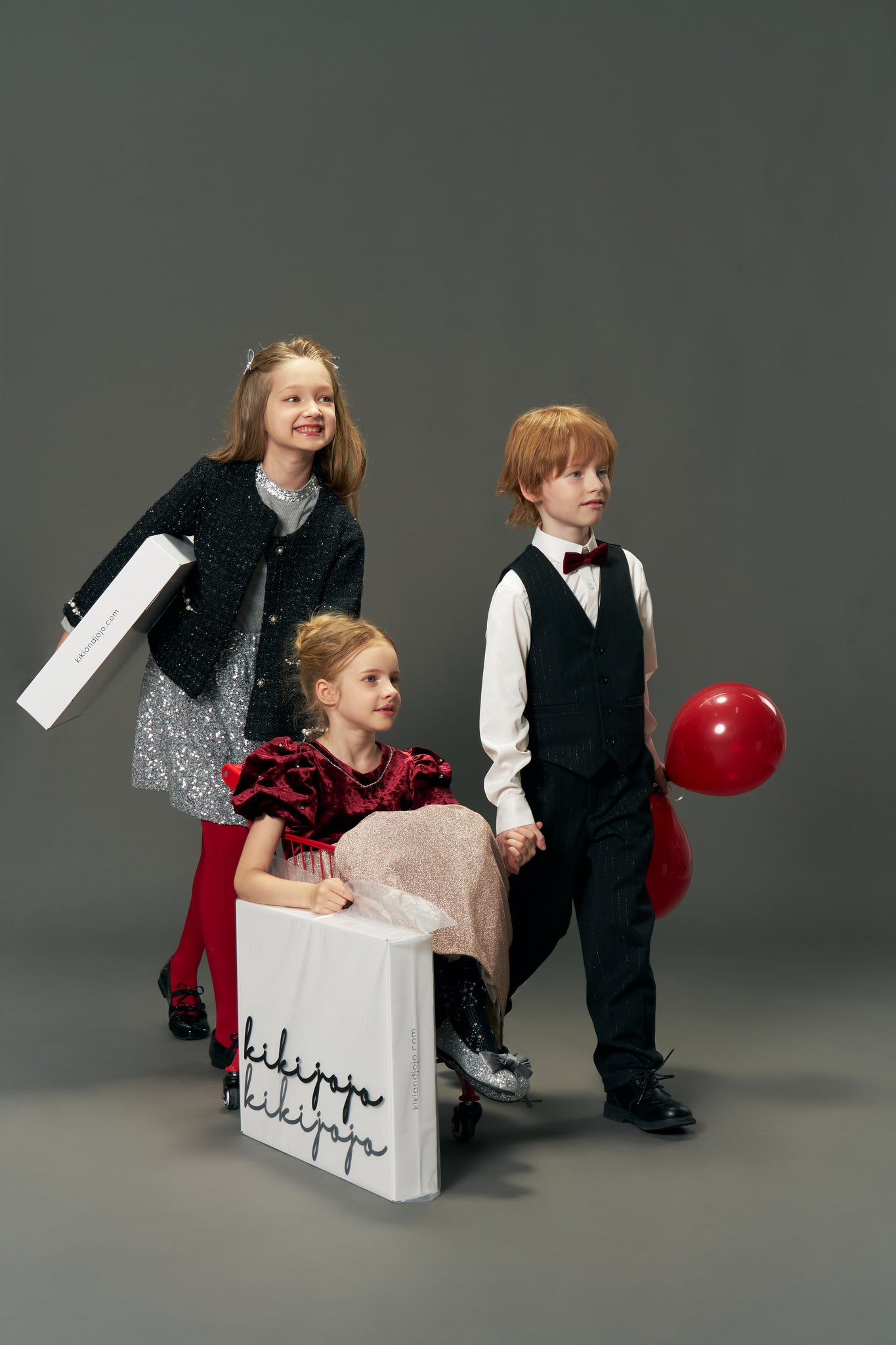 Three children in vintage-inspired luxury: one girl wears the KIKI and JOJO Girls Tweed Jacket – Classic Black Pearl, sitting in a wheeled box; another girl pushes it, while a boy stands beside them holding a red balloon.