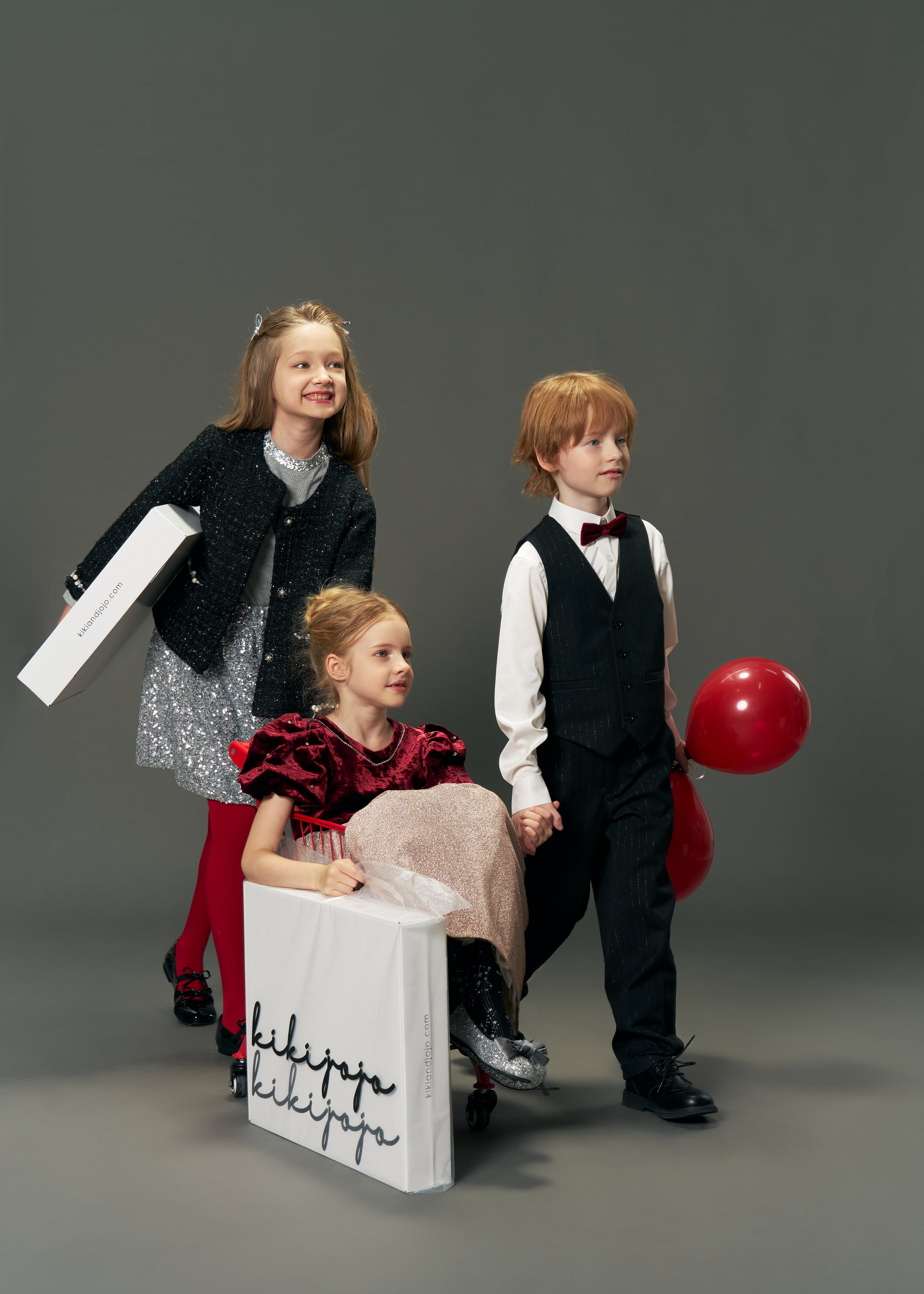 Three children in vintage-inspired luxury: one girl wears the KIKI and JOJO Girls Tweed Jacket – Classic Black Pearl, sitting in a wheeled box; another girl pushes it, while a boy stands beside them holding a red balloon.