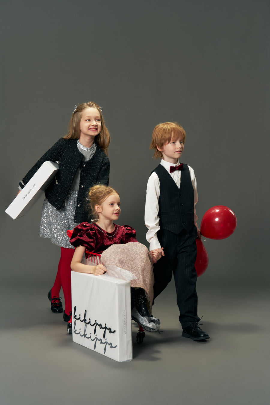 Three children in vintage-inspired luxury: one girl wears the KIKI and JOJO Girls Tweed Jacket – Classic Black Pearl, sitting in a wheeled box; another girl pushes it, while a boy stands beside them holding a red balloon.