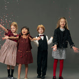 Four children in festive outfits stand smiling as confetti falls. One wears the KIKI and JOJO Girls Tweed Jacket – Classic Black Pearl, a vintage-inspired luxury piece with pearl details, set against a plain gray background.