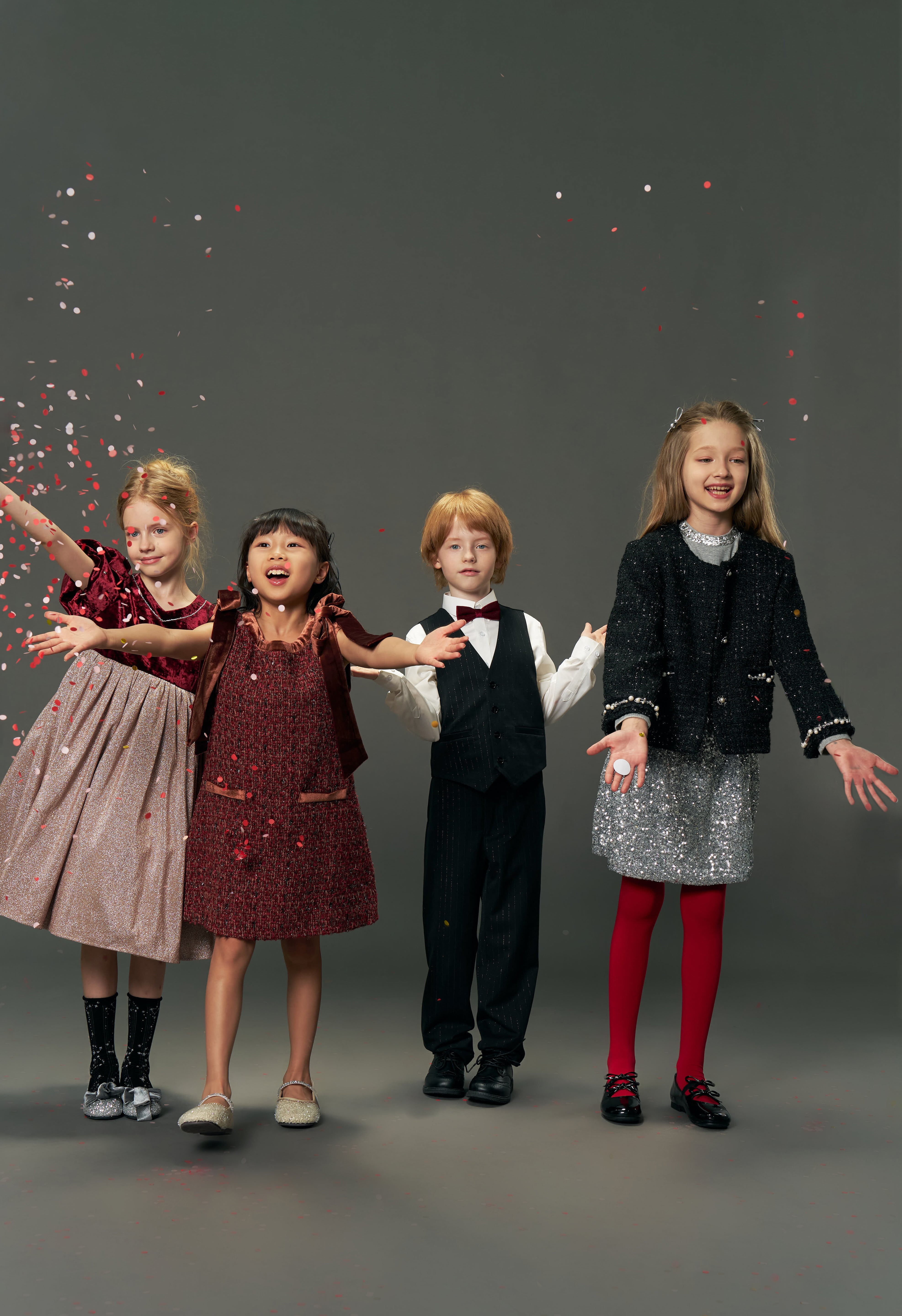 Four children in festive outfits stand smiling as confetti falls. One wears the KIKI and JOJO Girls Tweed Jacket – Classic Black Pearl, a vintage-inspired luxury piece with pearl details, set against a plain gray background.