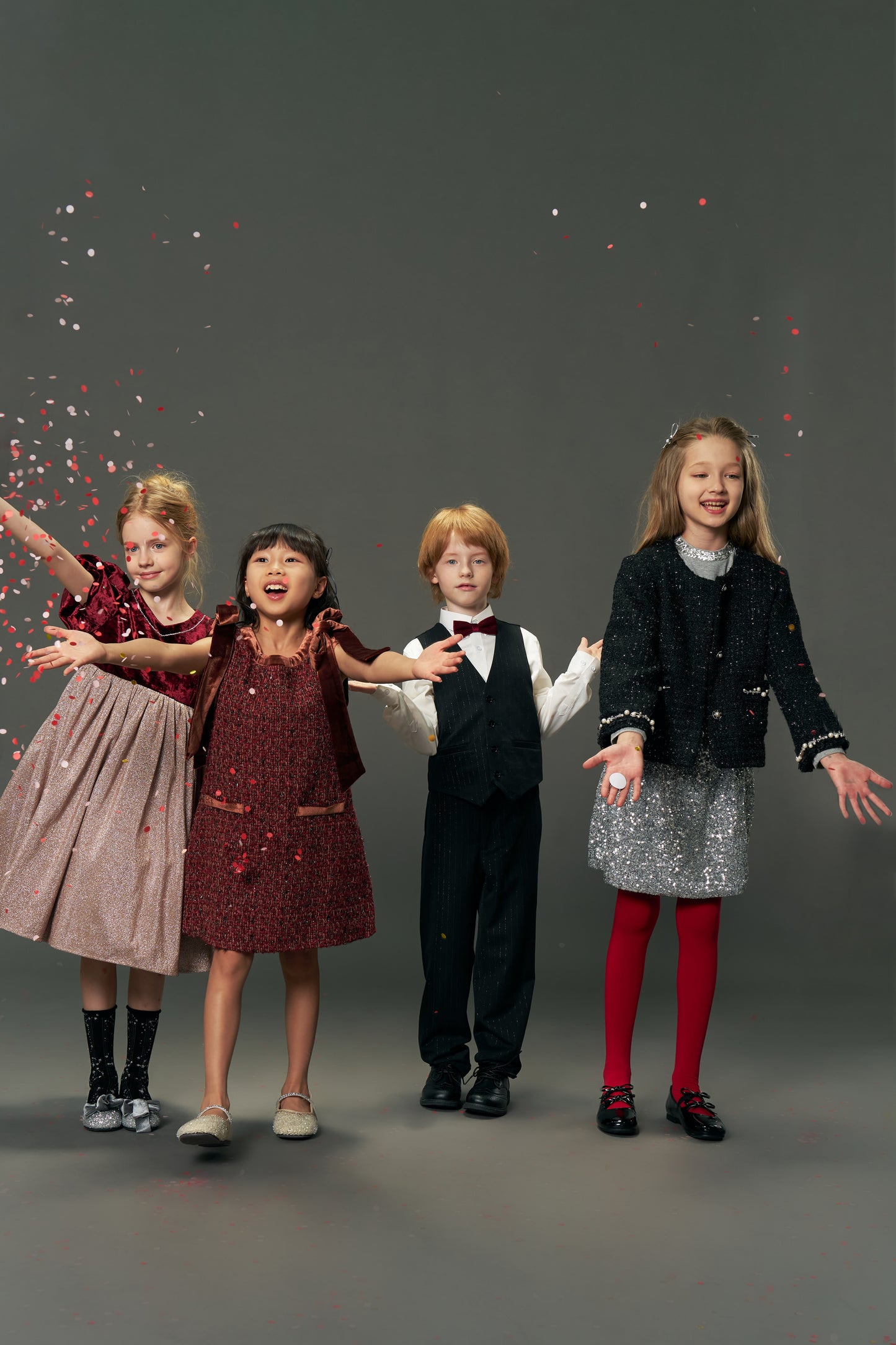 Four children in festive outfits stand smiling as confetti falls. One wears the KIKI and JOJO Girls Tweed Jacket – Classic Black Pearl, a vintage-inspired luxury piece with pearl details, set against a plain gray background.