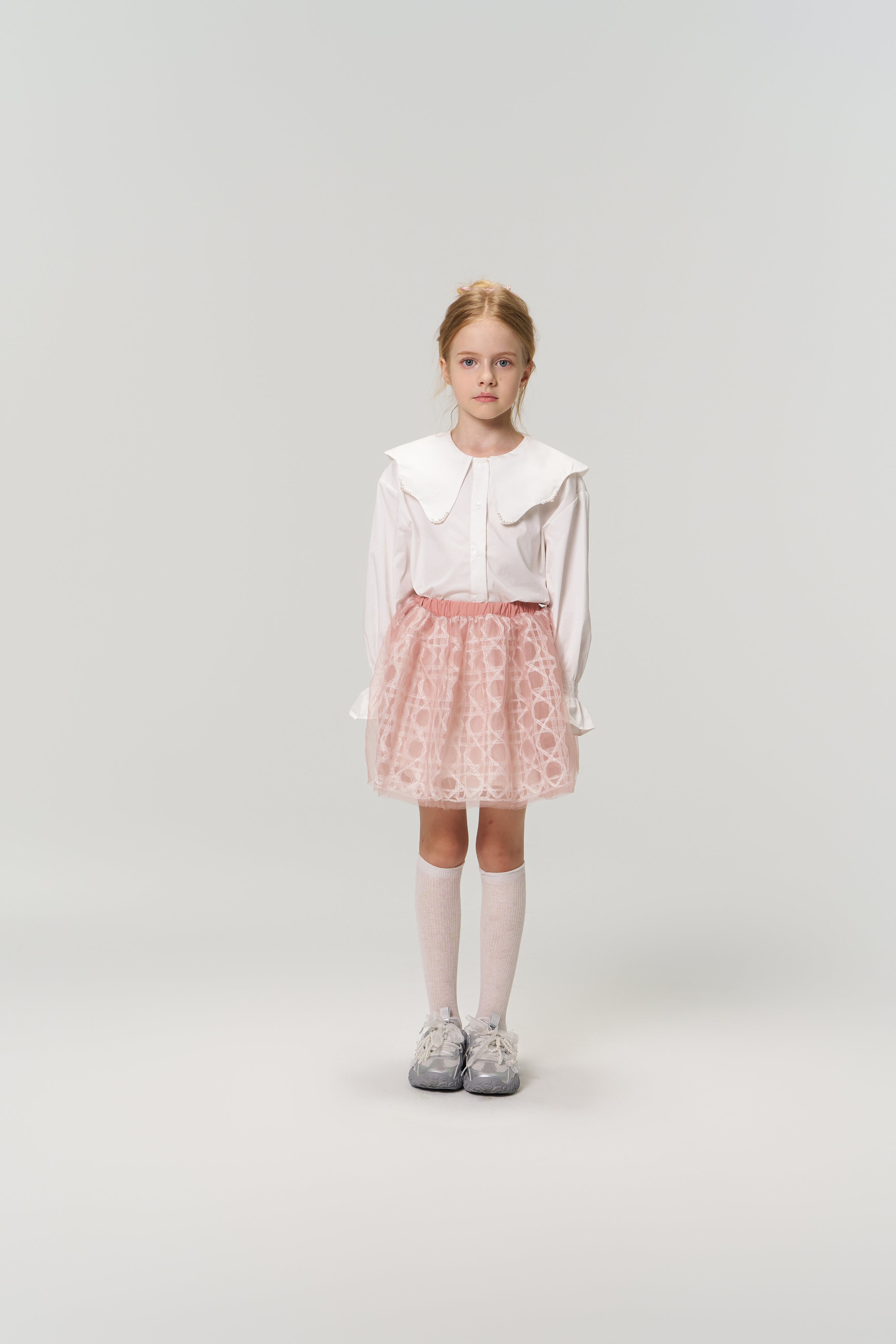 A young girl faces forward in the KIKI and JOJO Moonlit Whisper Blouse with a scalloped collar, paired with a pink skirt, white knee socks, and gray sneakers against a plain light background.