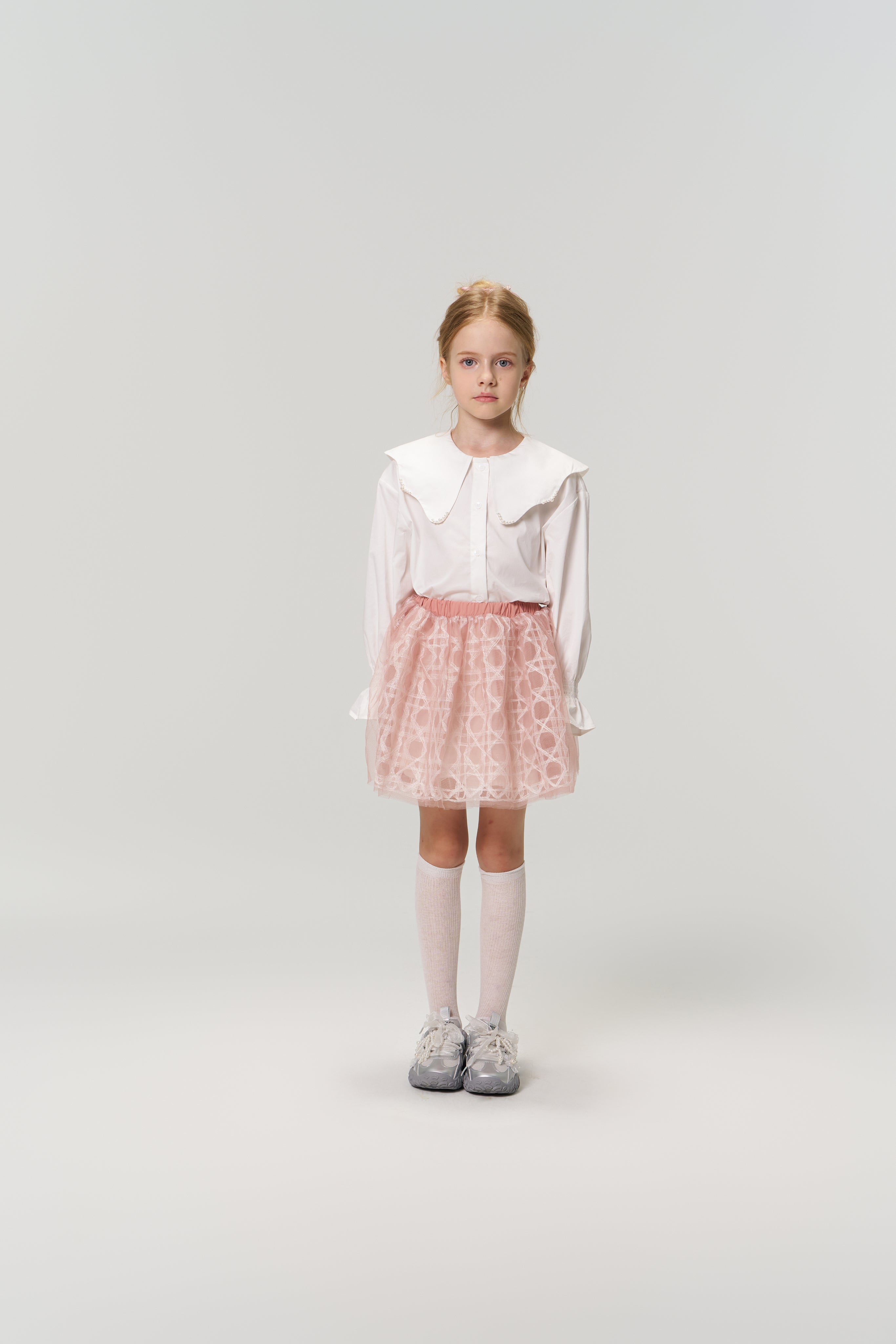 A young girl faces forward in the KIKI and JOJO Moonlit Whisper Blouse with a scalloped collar, paired with a pink skirt, white knee socks, and gray sneakers against a plain light background.