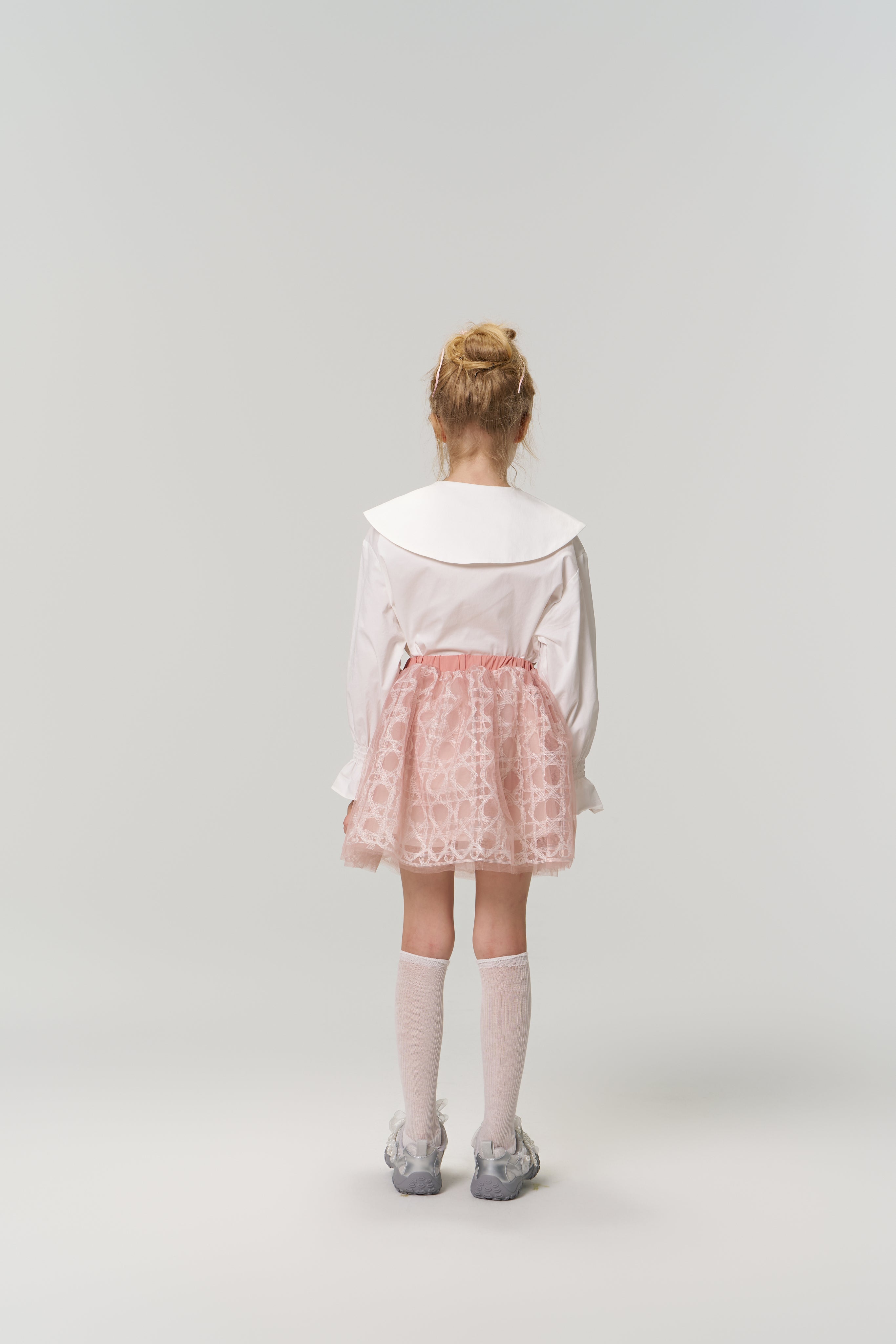 A young girl faces away, dressed in the KIKI and JOJO Moonlit Whisper Blouse—vintage style with puffed lantern sleeves and a scalloped collar—paired with a pink skirt, knee-high socks, and silver shoes against a plain light background.