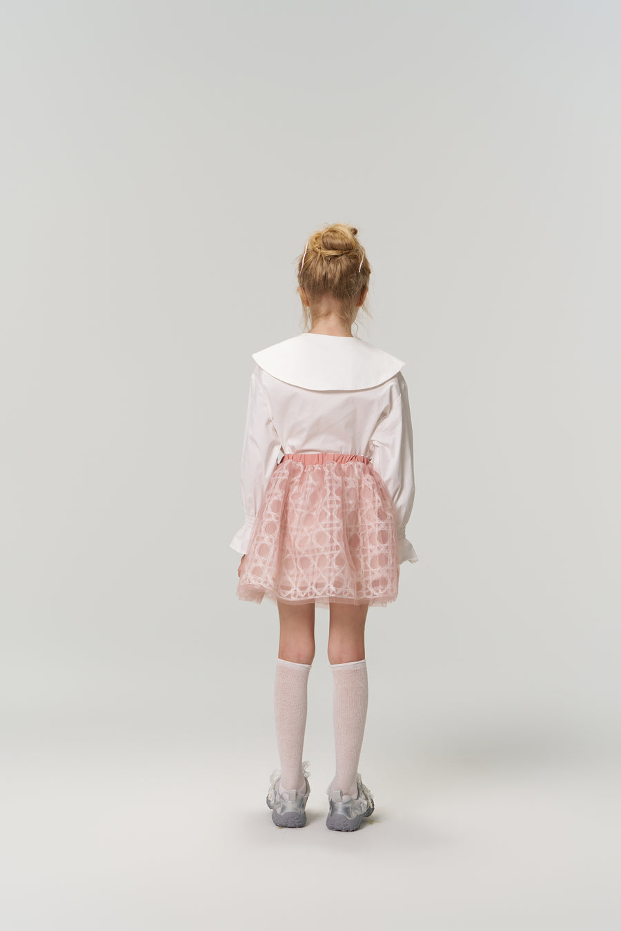 A young girl faces away, dressed in the KIKI and JOJO Moonlit Whisper Blouse—vintage style with puffed lantern sleeves and a scalloped collar—paired with a pink skirt, knee-high socks, and silver shoes against a plain light background.