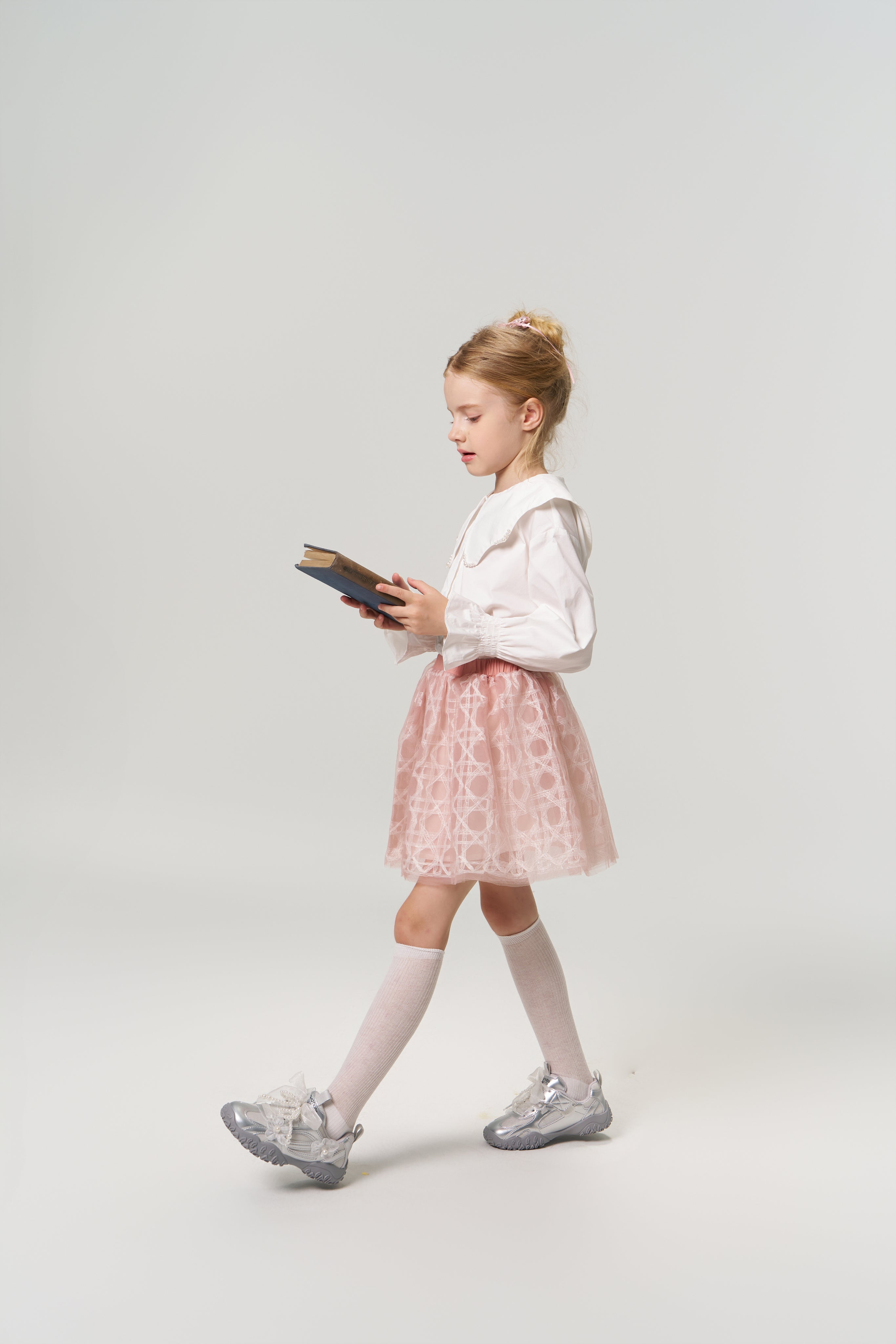 A young girl in the KIKI and JOJO Moonlit Whisper Blouse, featuring puffed lantern sleeves and a scalloped collar, pairs it with a pink skirt as she walks while reading against a plain light background.