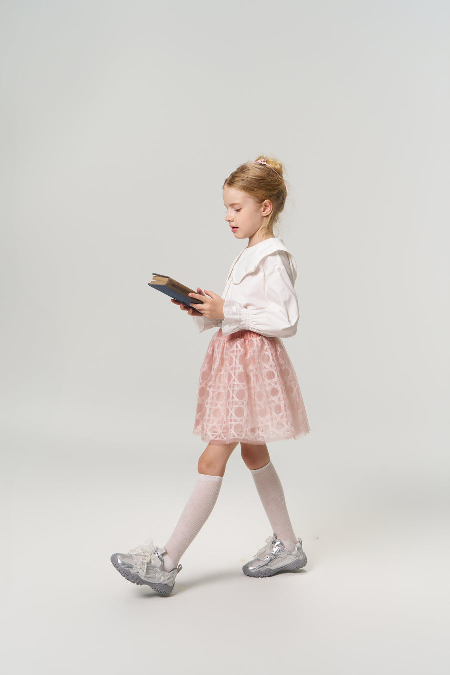 A young girl in the KIKI and JOJO Moonlit Whisper Blouse, featuring puffed lantern sleeves and a scalloped collar, pairs it with a pink skirt as she walks while reading against a plain light background.