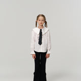 A young girl with braided hair faces forward, wearing KIKI and JOJO Petal Whisper Dress Pants with an elastic waistband, a white collared blouse, black tie, and silver shoes against a plain light background.