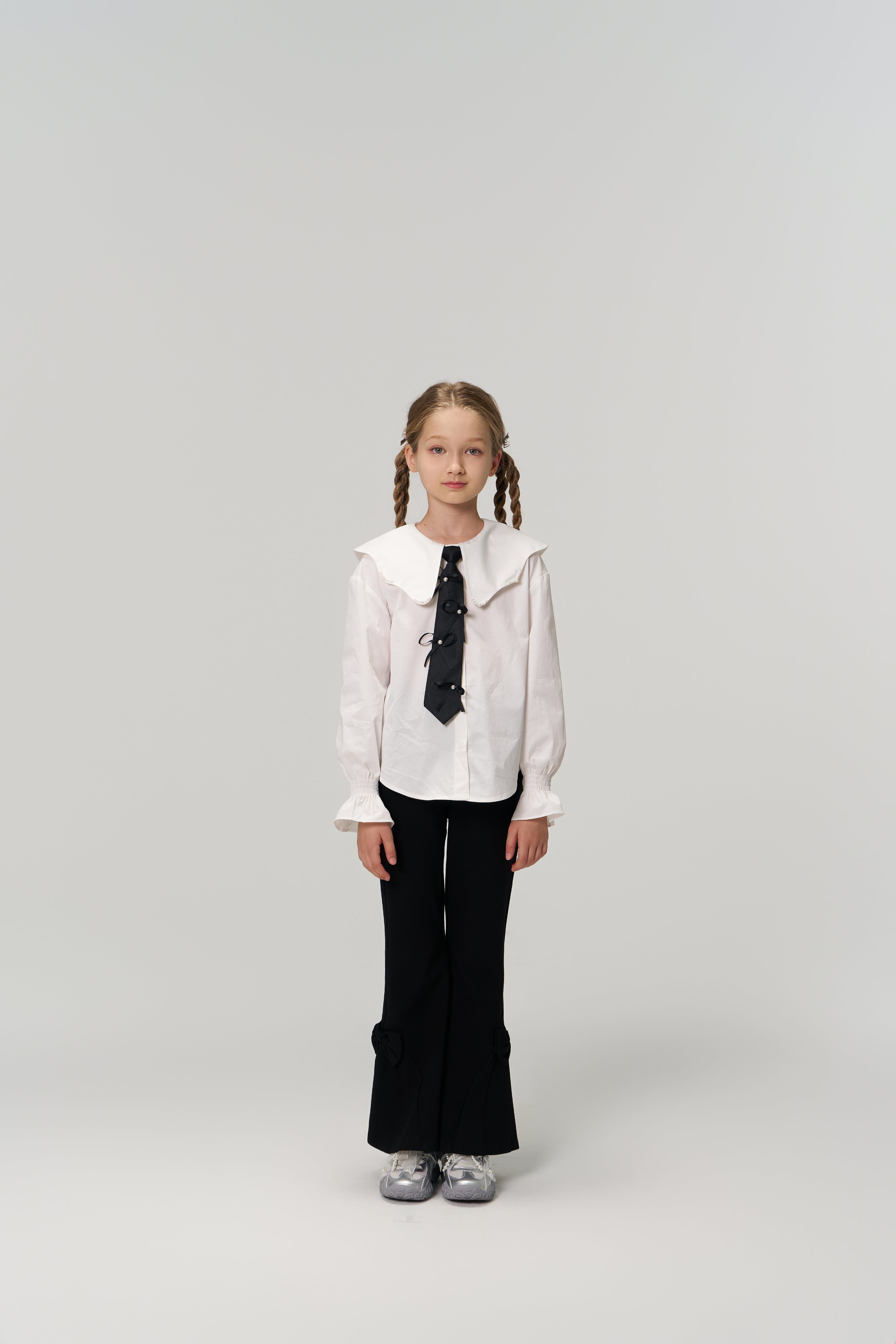 A young girl with braided hair faces forward, wearing KIKI and JOJO Petal Whisper Dress Pants with an elastic waistband, a white collared blouse, black tie, and silver shoes against a plain light background.