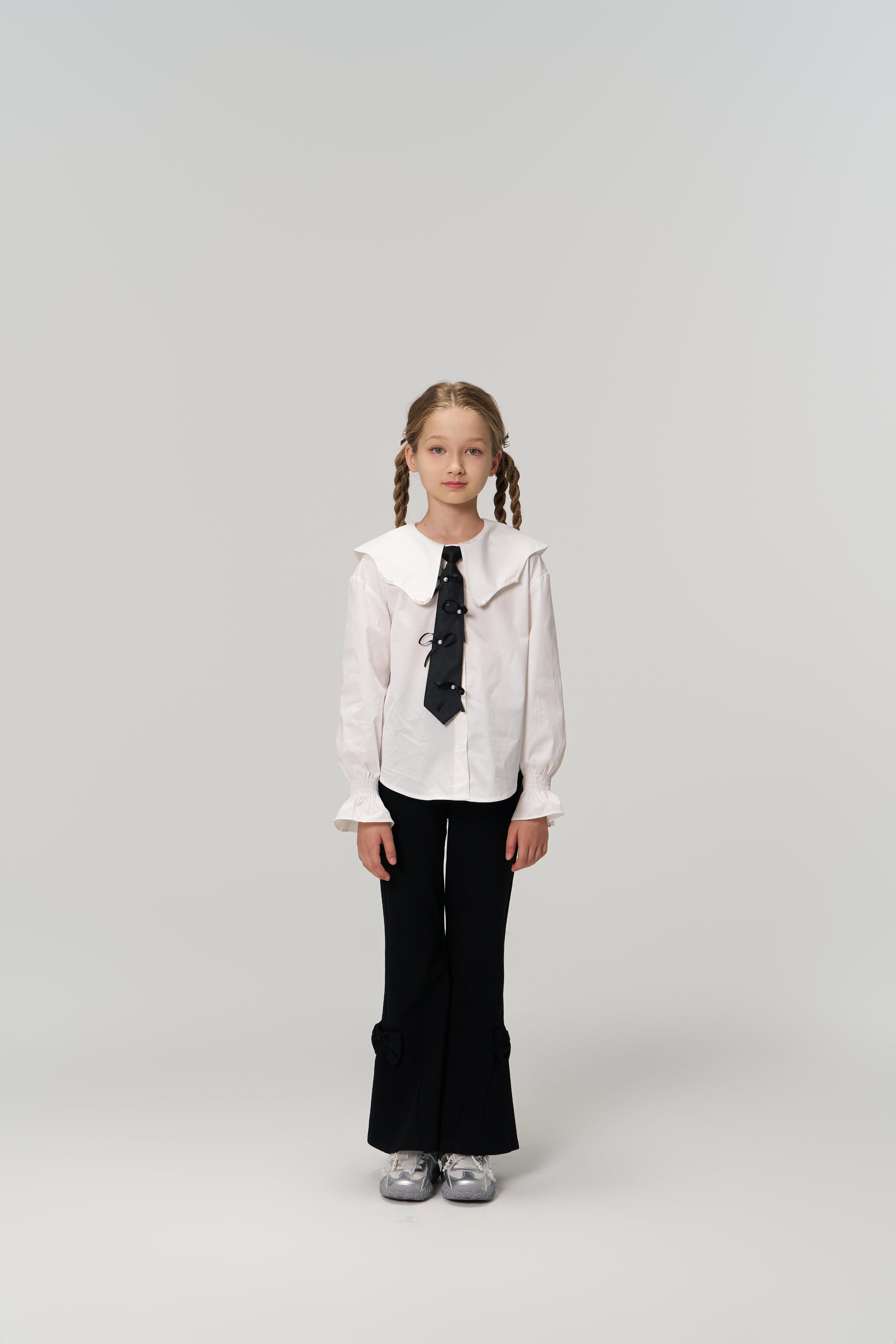 A young girl with braided hair faces forward, wearing KIKI and JOJO Petal Whisper Dress Pants with an elastic waistband, a white collared blouse, black tie, and silver shoes against a plain light background.