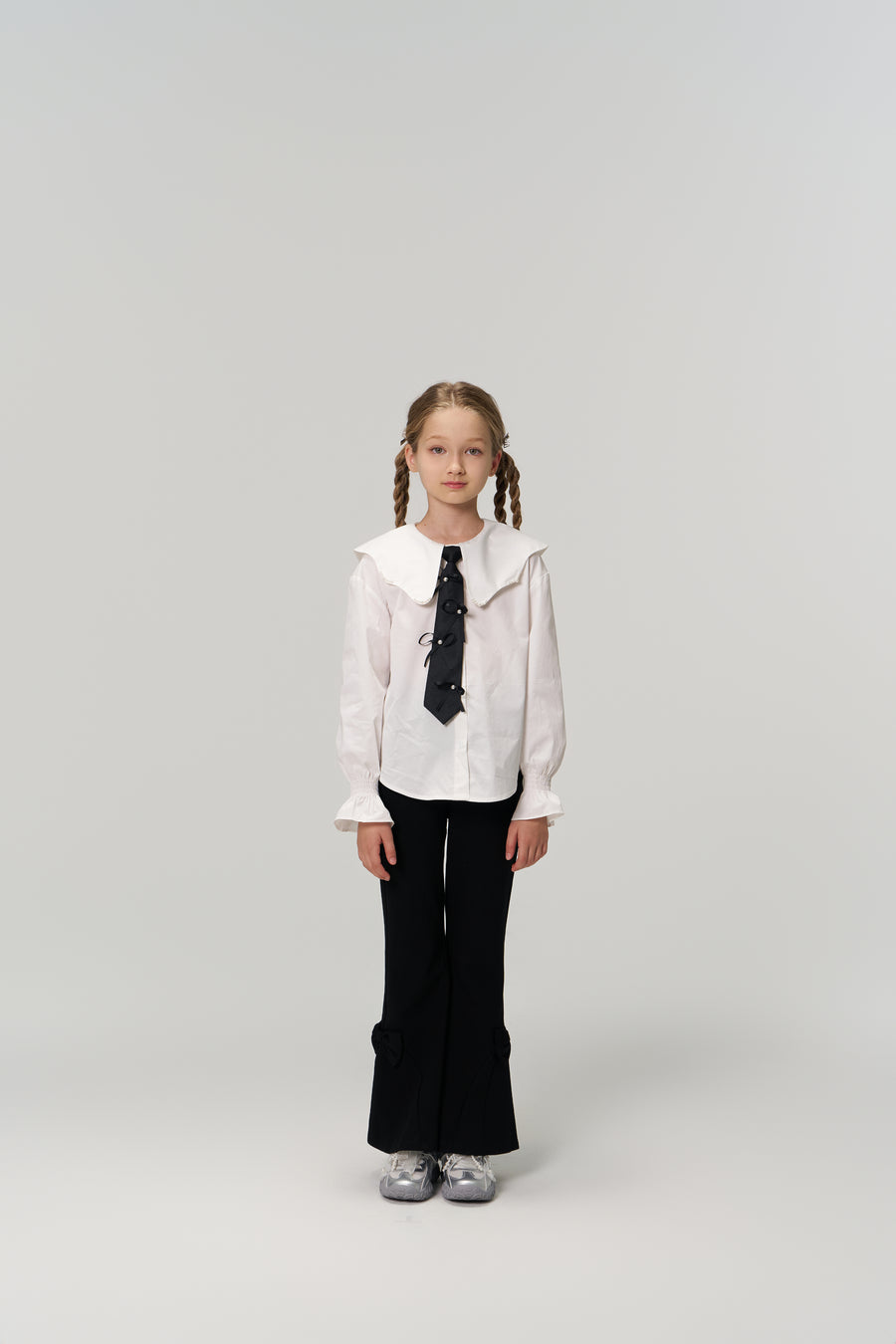 A young girl with braided hair faces forward, wearing KIKI and JOJO Petal Whisper Dress Pants with an elastic waistband, a white collared blouse, black tie, and silver shoes against a plain light background.