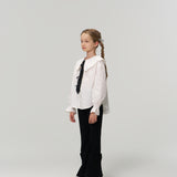 A young girl with braided hair stands against a plain background, wearing a white blouse, silver shoes, and KIKI and JOJO’s Petal Whisper Dress Pants—flared black pants with an elastic waistband.