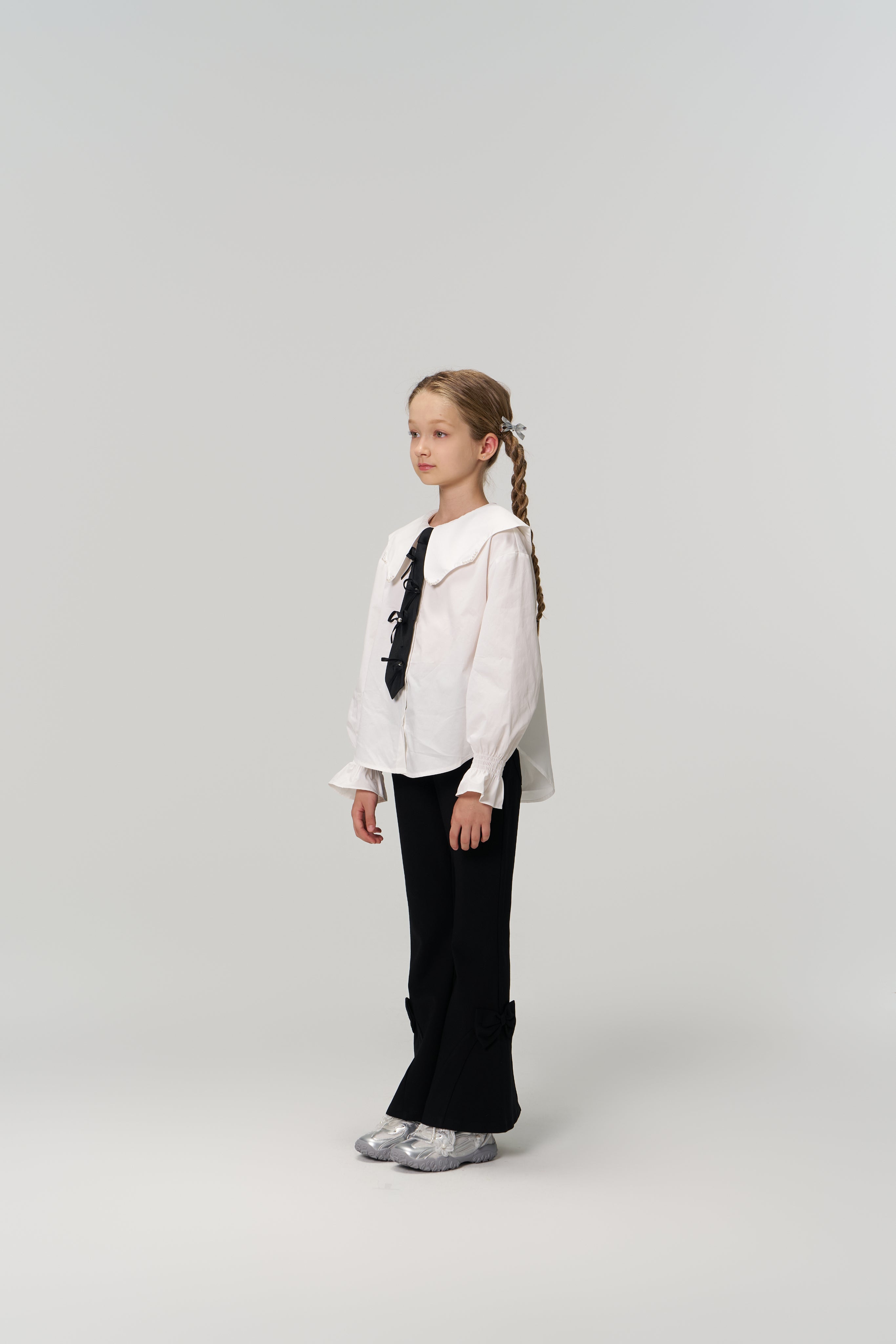 A young girl with braided hair stands against a plain background, wearing a white blouse, silver shoes, and KIKI and JOJO’s Petal Whisper Dress Pants—flared black pants with an elastic waistband.
