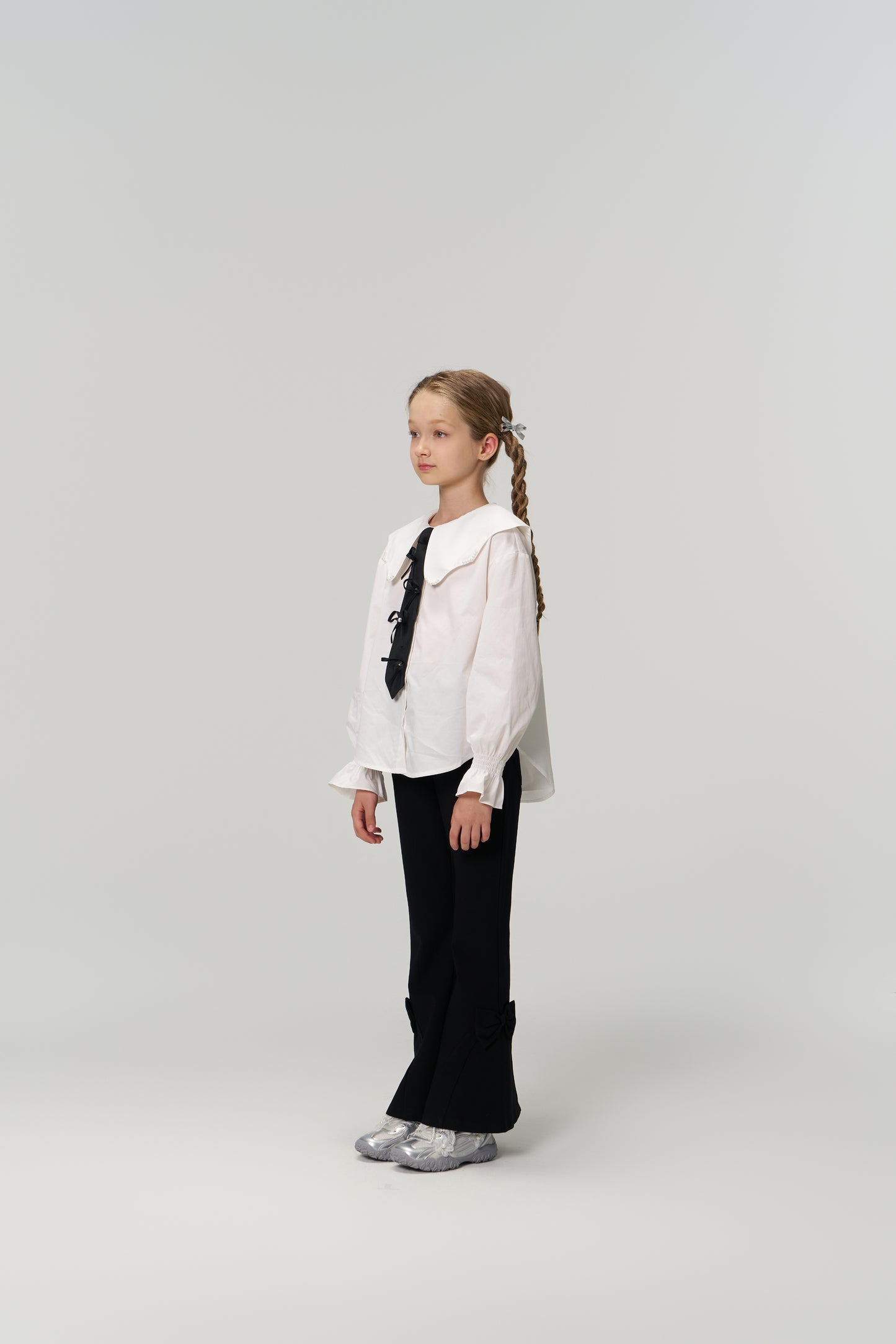 A young girl with braided hair stands against a plain background, wearing a white blouse, silver shoes, and KIKI and JOJO’s Petal Whisper Dress Pants—flared black pants with an elastic waistband.