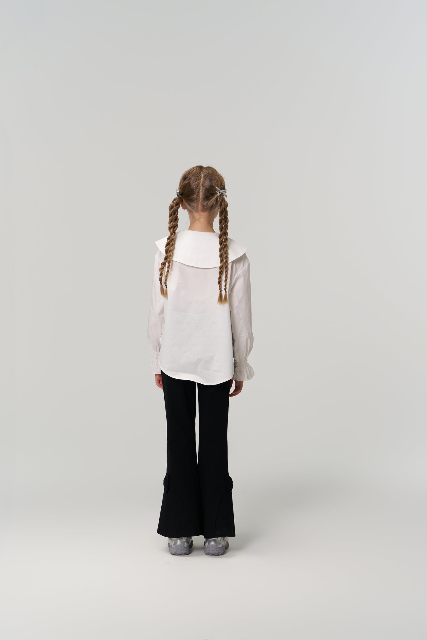 A young girl with braided hair faces away, wearing KIKI and JOJO's Petal Whisper Dress Pants—flared black with an elastic waistband—and a white blouse, set against a plain light background.