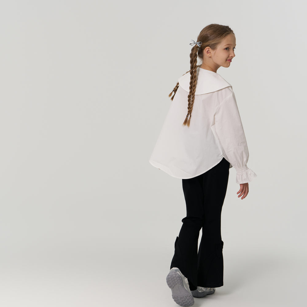 Young girl with braided hair, wearing a white blouse and black pants, walking away and turning her head to the side in a plain, light-colored studio setting.