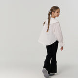 Young girl with braided hair, wearing a white blouse and black pants, walking away and turning her head to the side in a plain, light-colored studio setting.