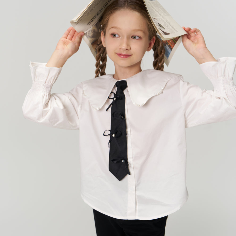 Young girl with braided hair holds a newspaper over her head like a hat, wearing a white blouse and black tie, standing against a plain light background.