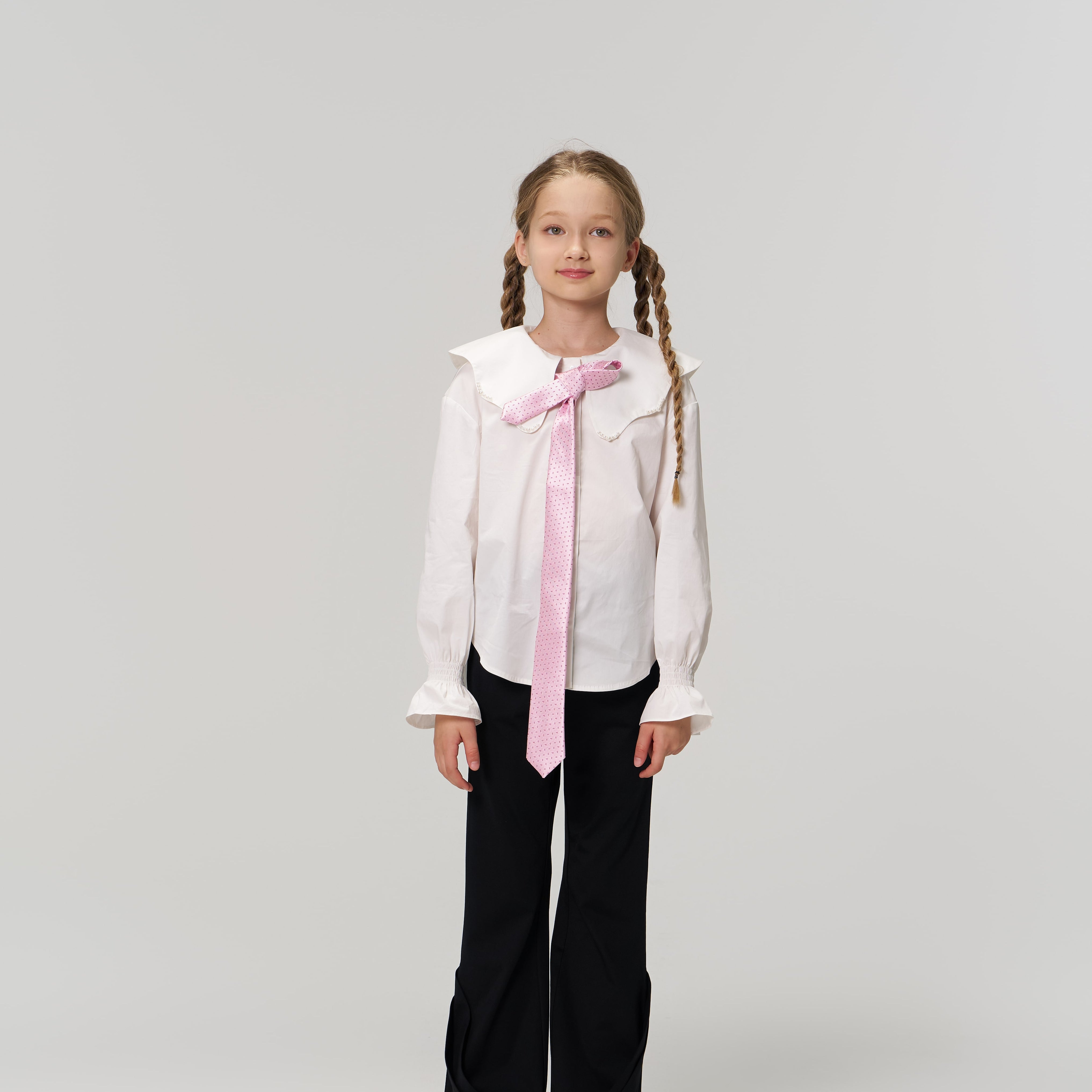 A young girl with braided hair faces forward, wearing a white blouse, pink tie, and KIKI and JOJO Echo Whisper Dress Pants with an elastic waistband, paired with gray sneakers against a plain light background.