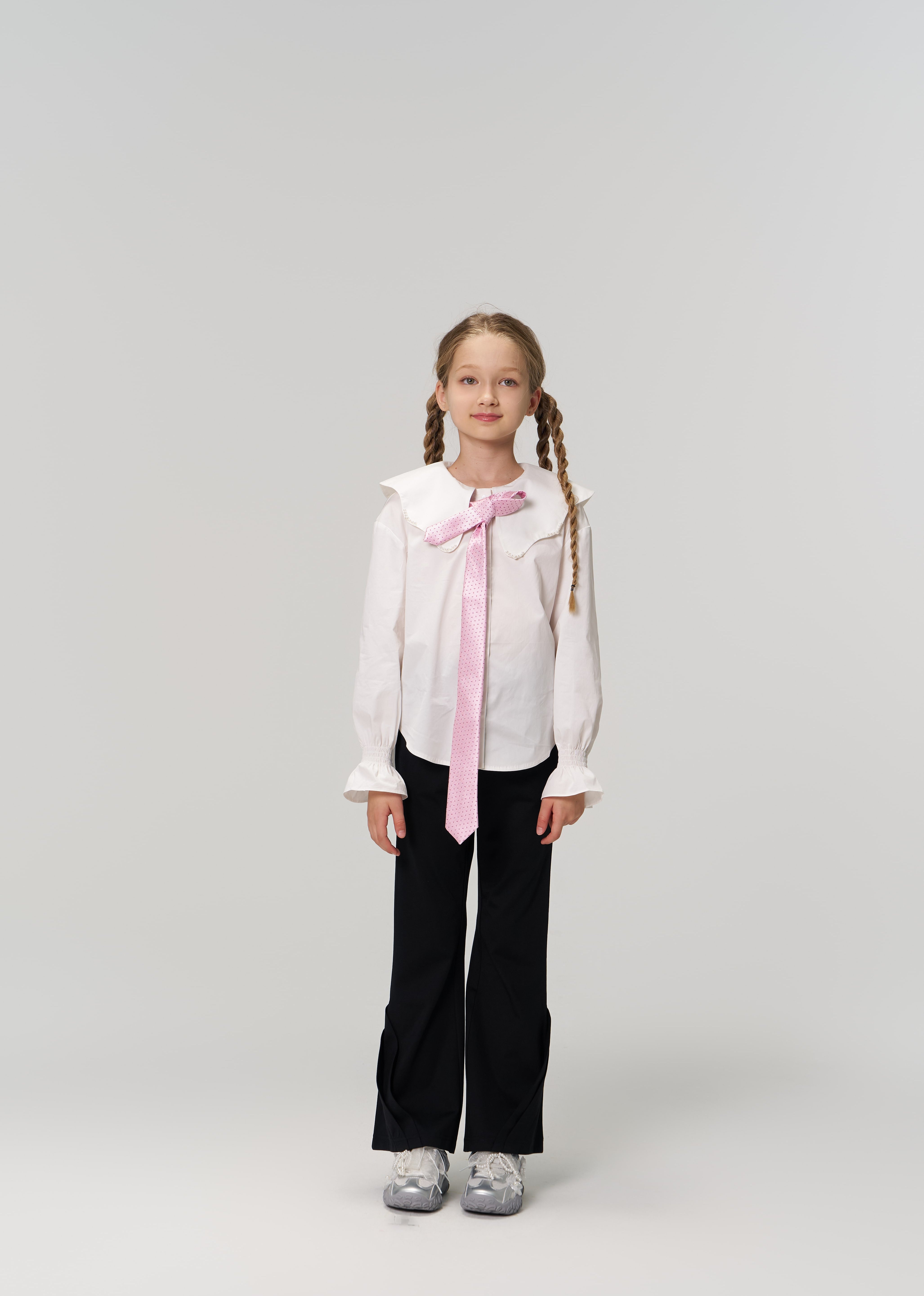 A young girl with braided hair faces forward, wearing a white blouse, pink tie, and KIKI and JOJO Echo Whisper Dress Pants with an elastic waistband, paired with gray sneakers against a plain light background.