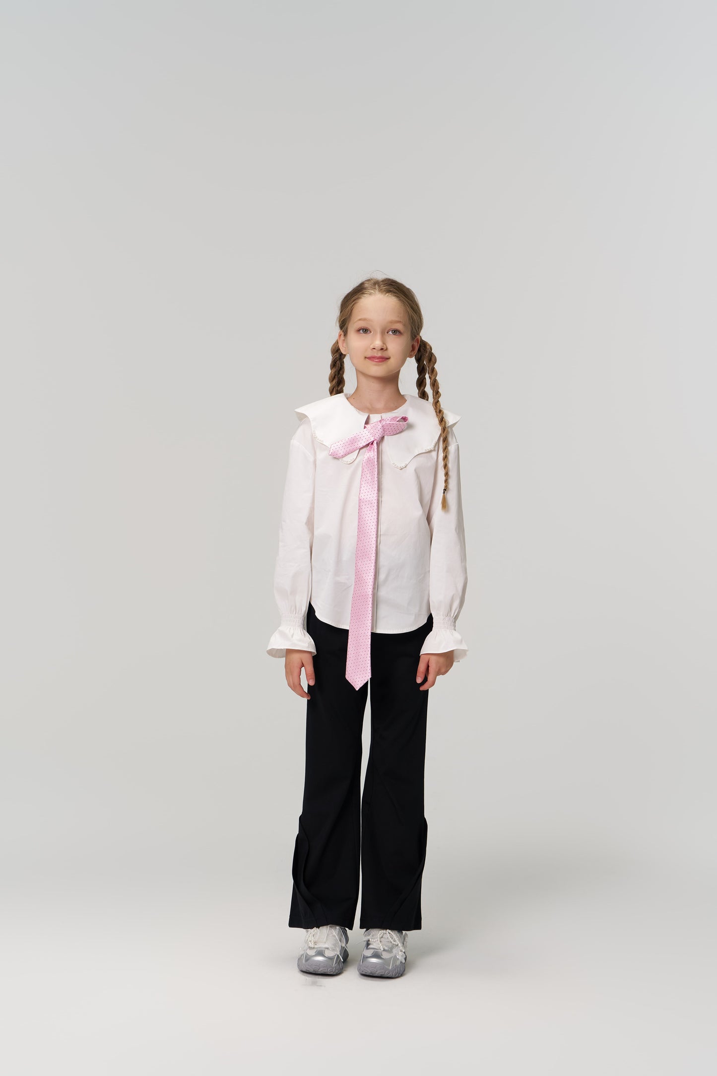 A young girl with braided hair faces forward, wearing a white blouse, pink tie, and KIKI and JOJO Echo Whisper Dress Pants with an elastic waistband, paired with gray sneakers against a plain light background.