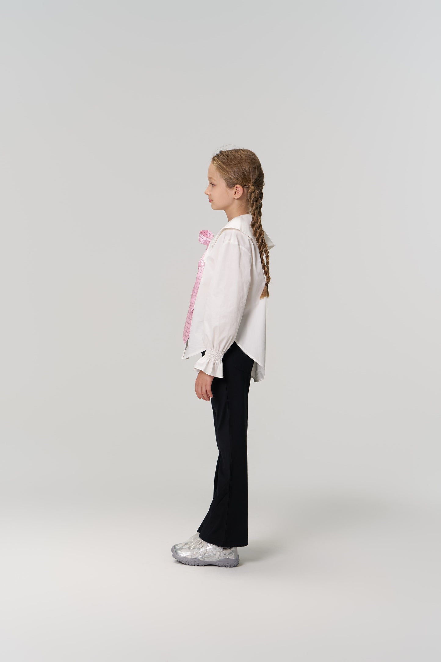 A young girl with braided hair, wearing a white shirt, KIKI and JOJO Echo Whisper Dress Pants with an elastic waistband, and silver shoes stands in profile against a plain light background.