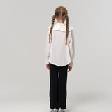 A young girl with braided hair faces away, wearing a white blouse and KIKI and JOJO's Echo Whisper Dress Pants—flared black pants with an elastic waistband—paired with silver shoes against a plain light background.