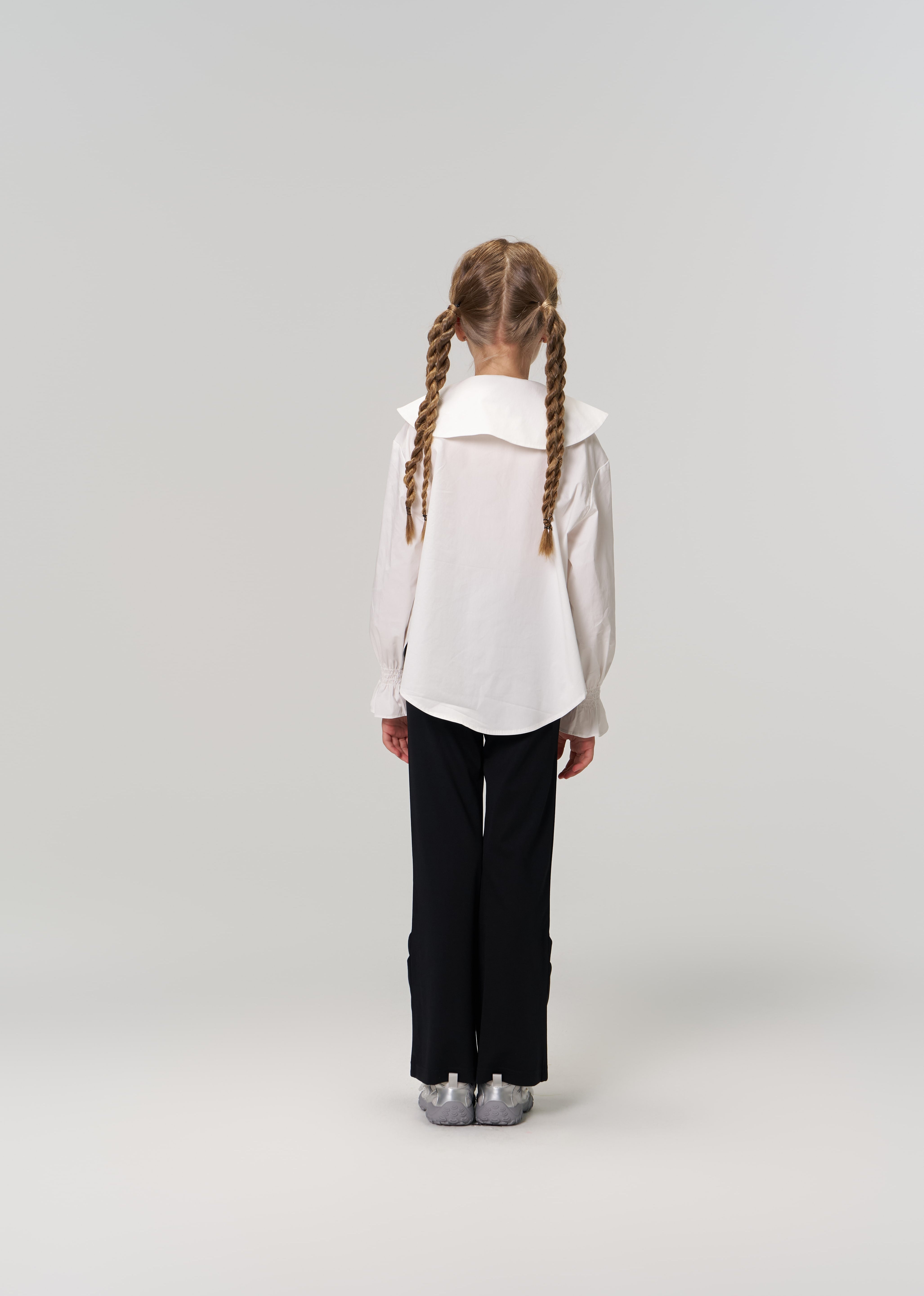A young girl with braided hair faces away, wearing a white blouse and KIKI and JOJO's Echo Whisper Dress Pants—flared black pants with an elastic waistband—paired with silver shoes against a plain light background.