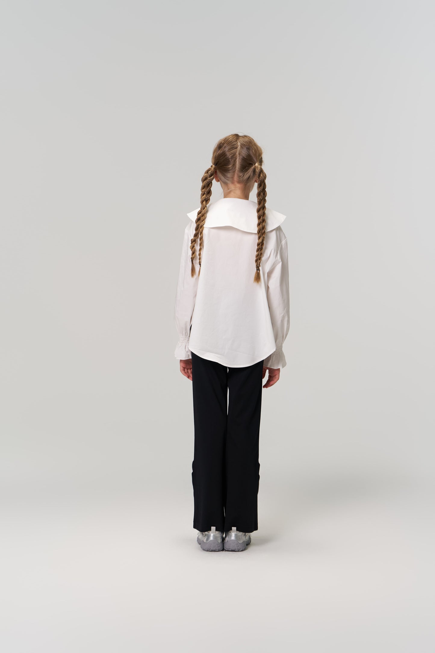 A young girl with braided hair faces away, wearing a white blouse and KIKI and JOJO's Echo Whisper Dress Pants—flared black pants with an elastic waistband—paired with silver shoes against a plain light background.