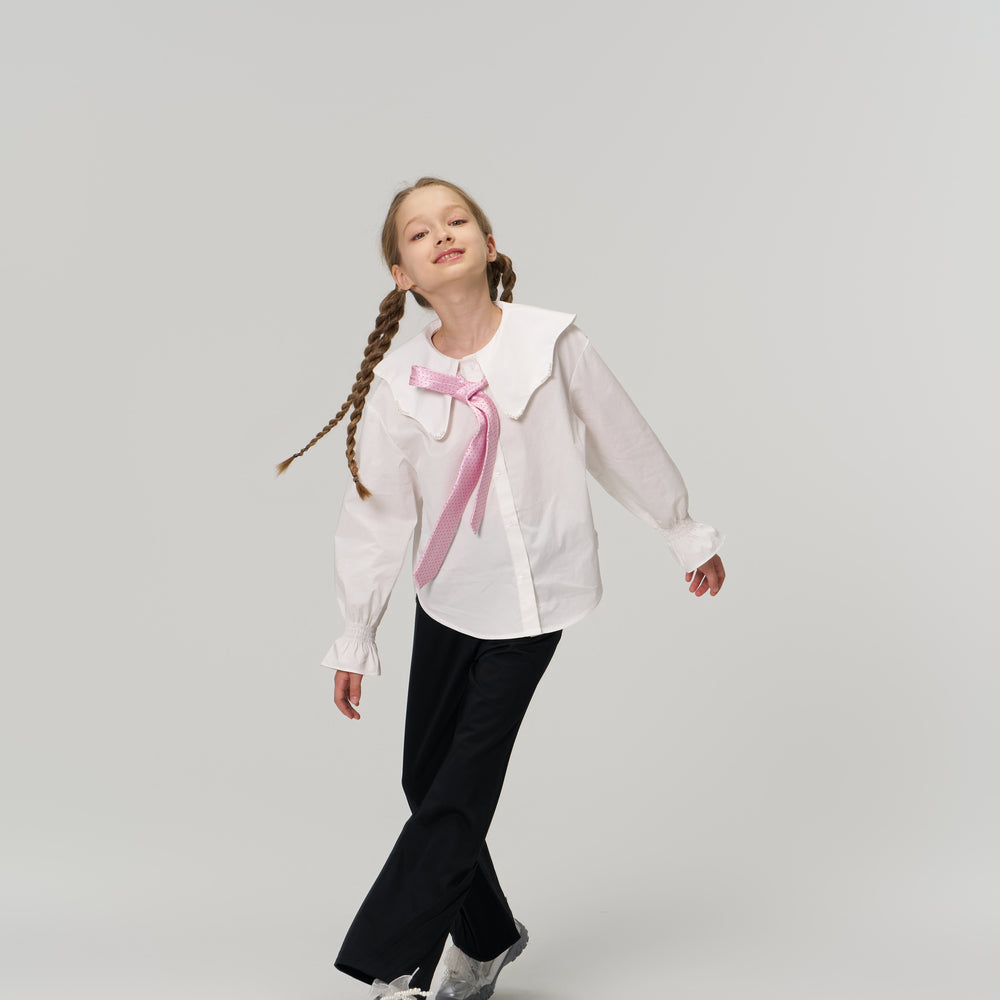 Young girl with braided hair walking, wearing a white blouse with a large collar and pink ribbon, black pants, and sneakers, against a plain light background.