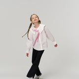 Young girl with braided hair walking, wearing a white blouse with a large collar and pink ribbon, black pants, and sneakers, against a plain light background.