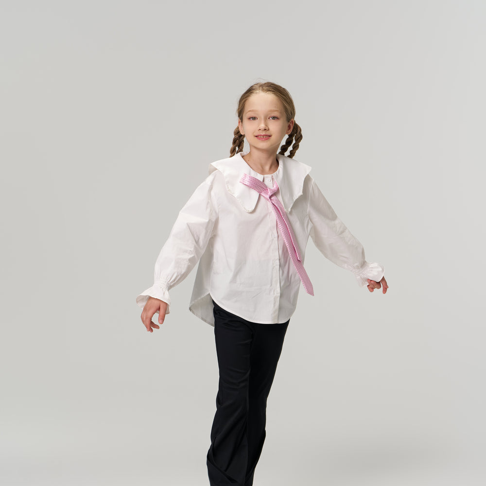 A young girl with braided hair wearing a white shirt, pink tie, black pants, and sneakers stands against a plain light background, arms slightly outstretched.