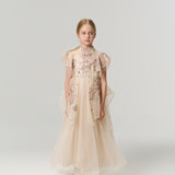 A young girl poses against a plain backdrop in the KIKI and JOJO Jade Whisper Dress, a long, light-hued cheongsam featuring embroidered florals, decorative sleeves, mandarin collar, and fluffy shoes.