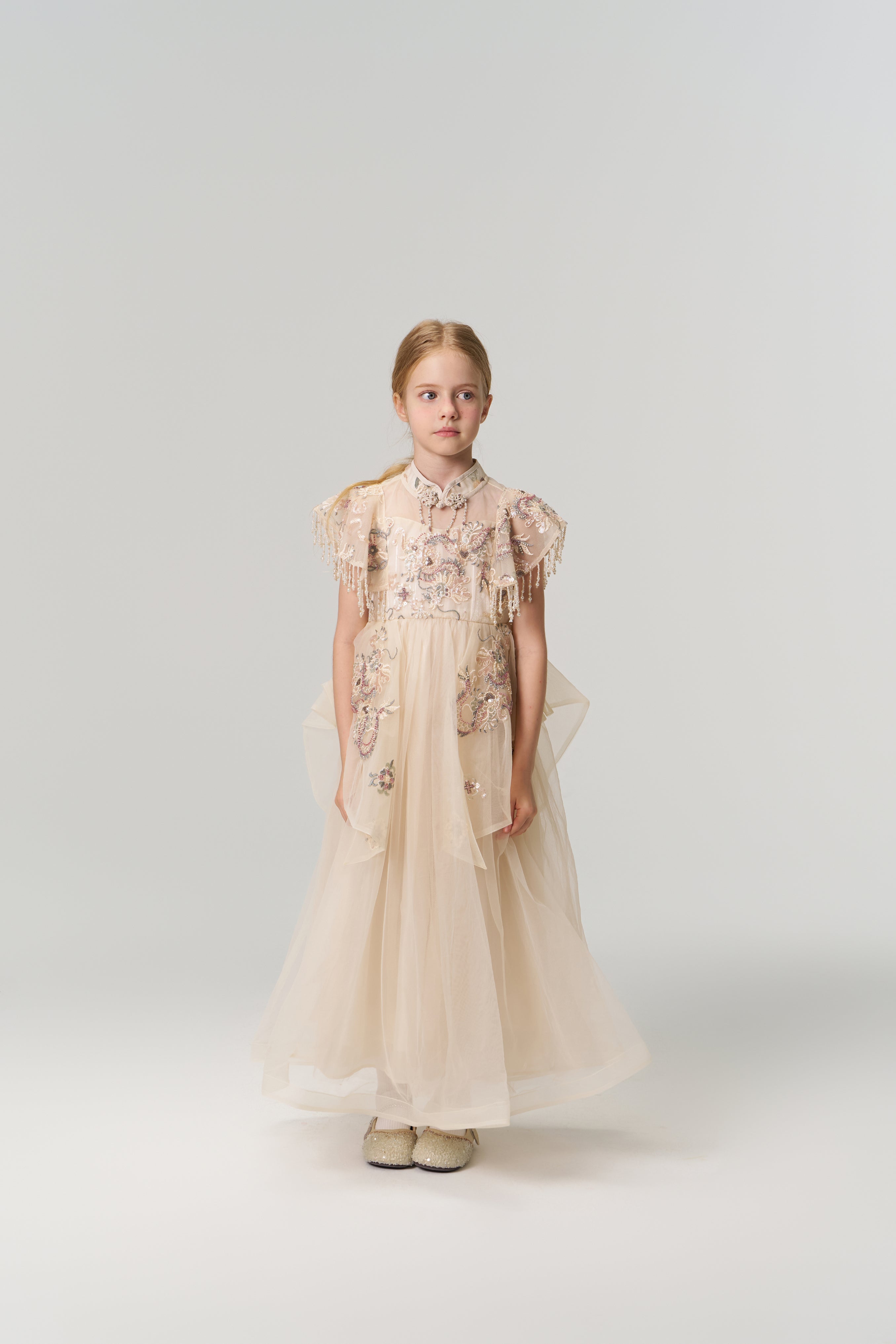 A young girl poses against a plain backdrop in the KIKI and JOJO Jade Whisper Dress, a long, light-hued cheongsam featuring embroidered florals, decorative sleeves, mandarin collar, and fluffy shoes.