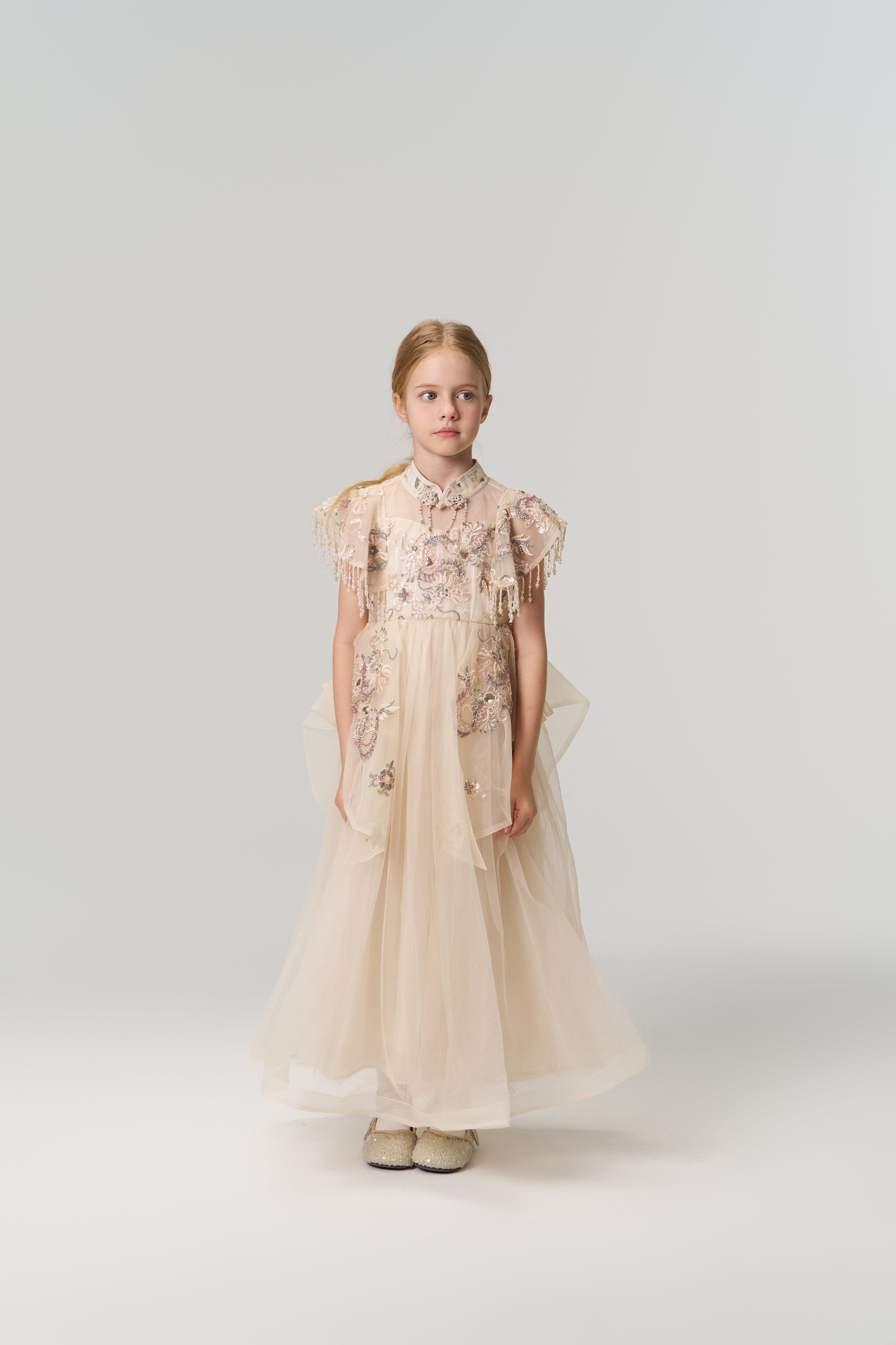 A young girl poses against a plain backdrop in the KIKI and JOJO Jade Whisper Dress, a long, light-hued cheongsam featuring embroidered florals, decorative sleeves, mandarin collar, and fluffy shoes.