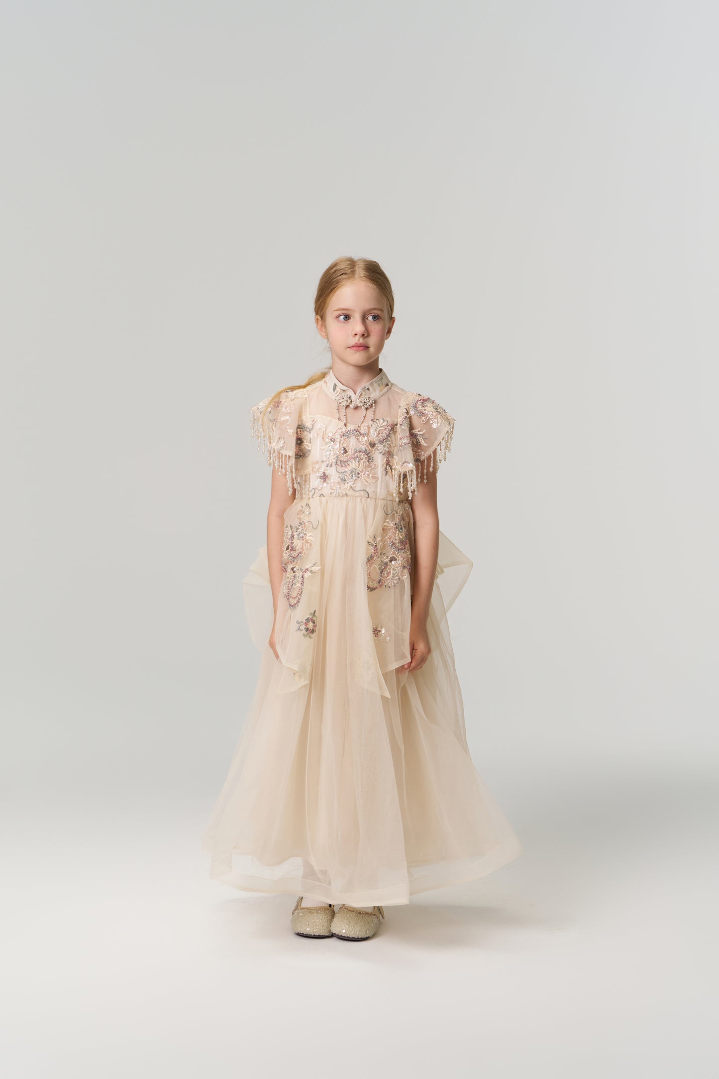 A young girl poses against a plain backdrop in the KIKI and JOJO Jade Whisper Dress, a long, light-hued cheongsam featuring embroidered florals, decorative sleeves, mandarin collar, and fluffy shoes.